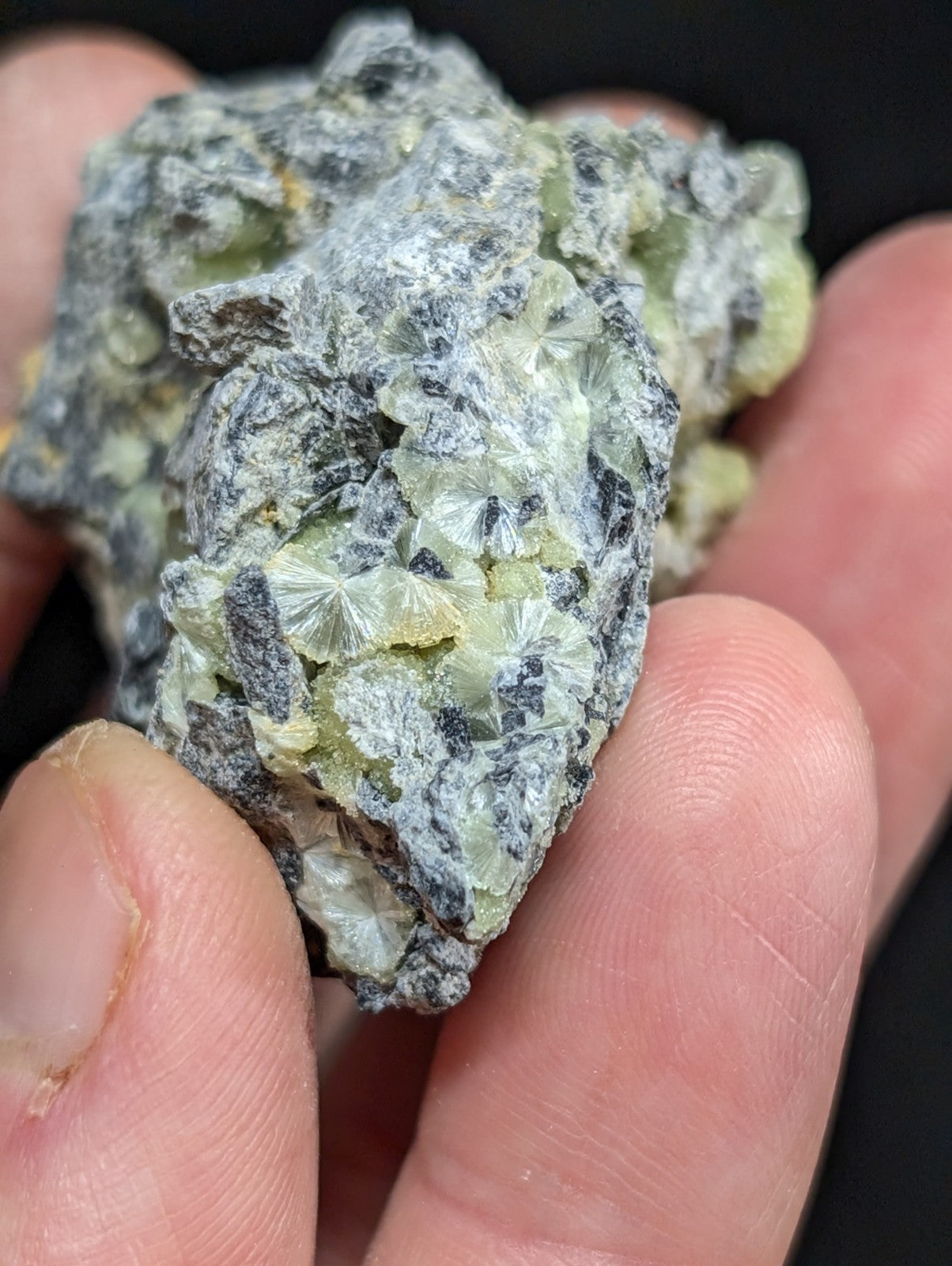 A hand holds a rough, irregularly shaped mineral specimen featuring translucent pale green crystals with radial facets embedded in a dark gray matrix.