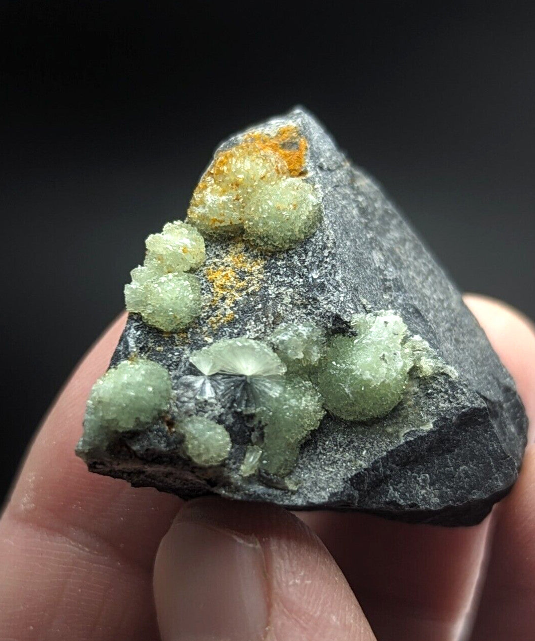 A hand holds a dark gray rock specimen featuring clusters of translucent green crystals and patches of orange mineral growth.