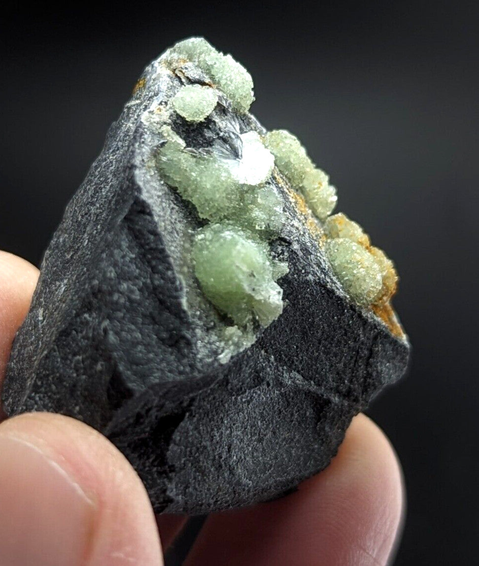 A dark, rough-textured rock specimen featuring clusters of translucent green crystals with hints of yellowish mineral growth.