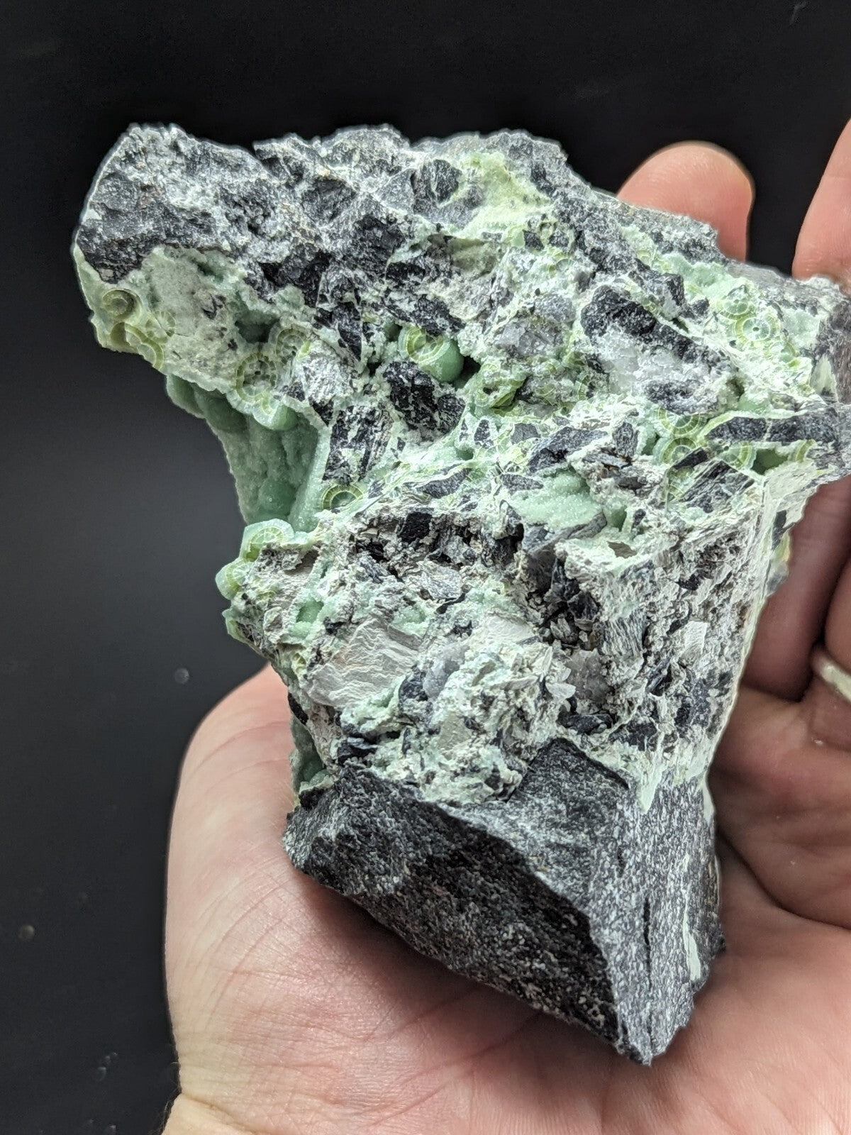 A hand holds a rough, irregularly shaped piece of green and black mineral rock with visible crystalline textures.
