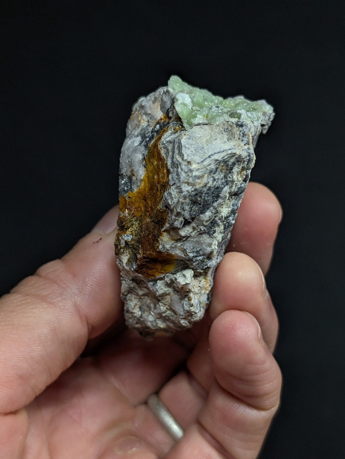 A hand holds a rough, irregularly shaped mineral specimen featuring a prominent orange-brown vein running through its center and patches of translucent green crystal at the top.