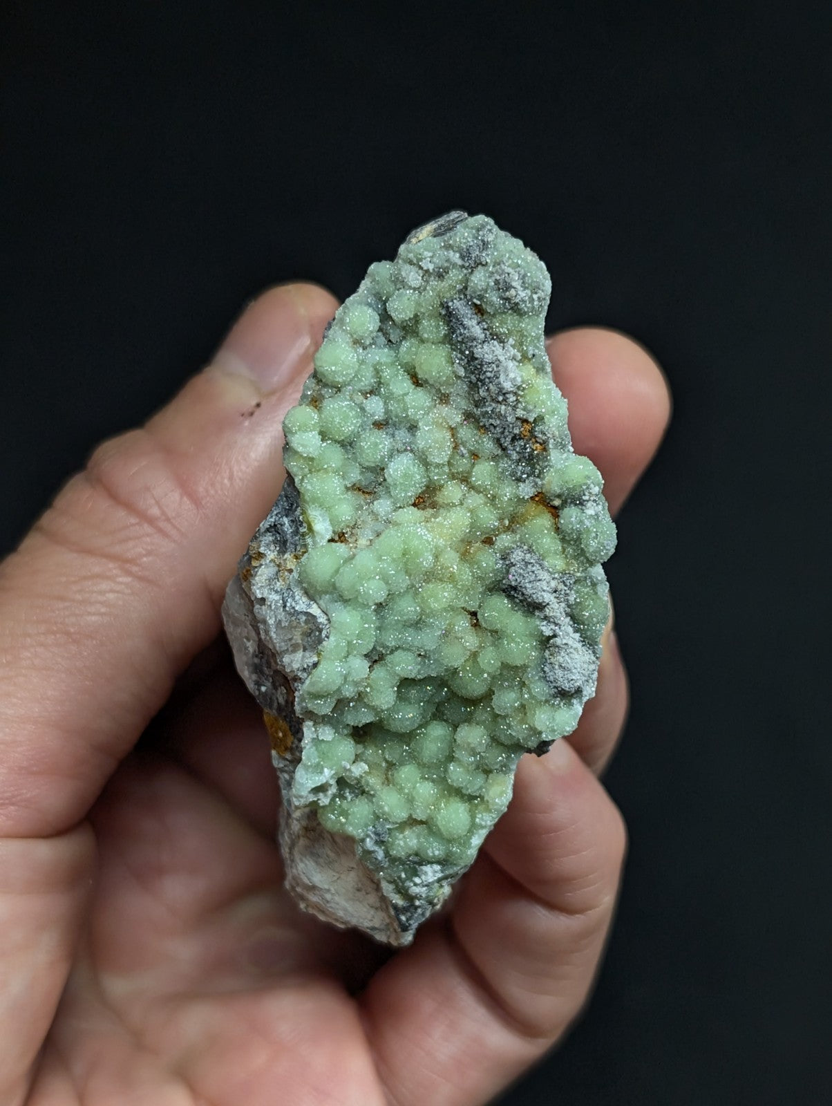 A hand holds a rough, irregularly shaped mineral specimen featuring clusters of pale green, rounded crystals growing from a darker gray matrix.
