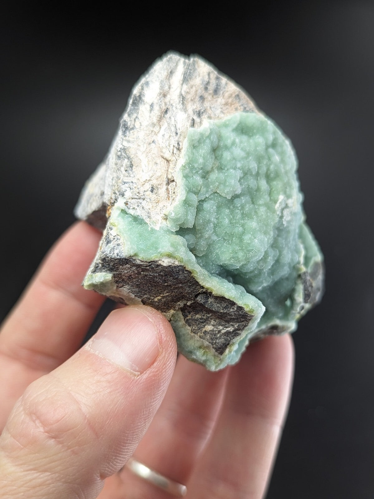 A hand holds a rough, irregularly shaped piece of green mineral with crystalline texture and white-gray matrix.