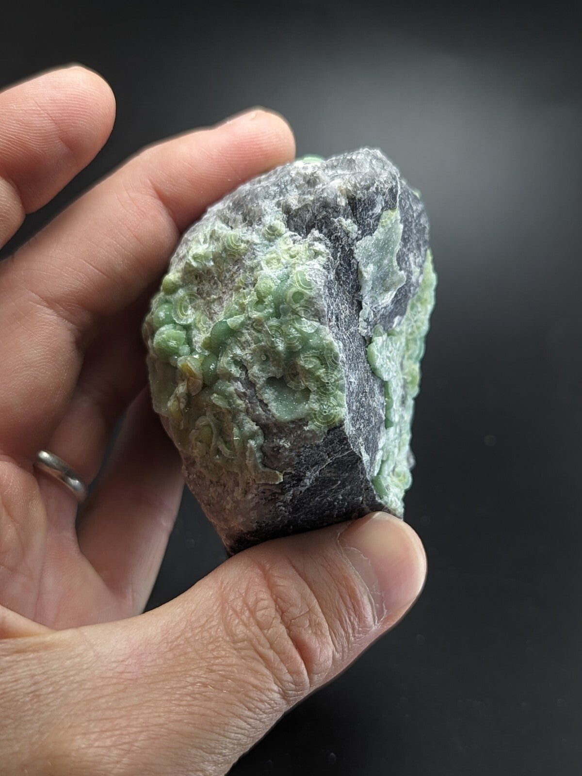 A hand holds a rough, irregularly shaped piece of green and gray mineral with textured surfaces.