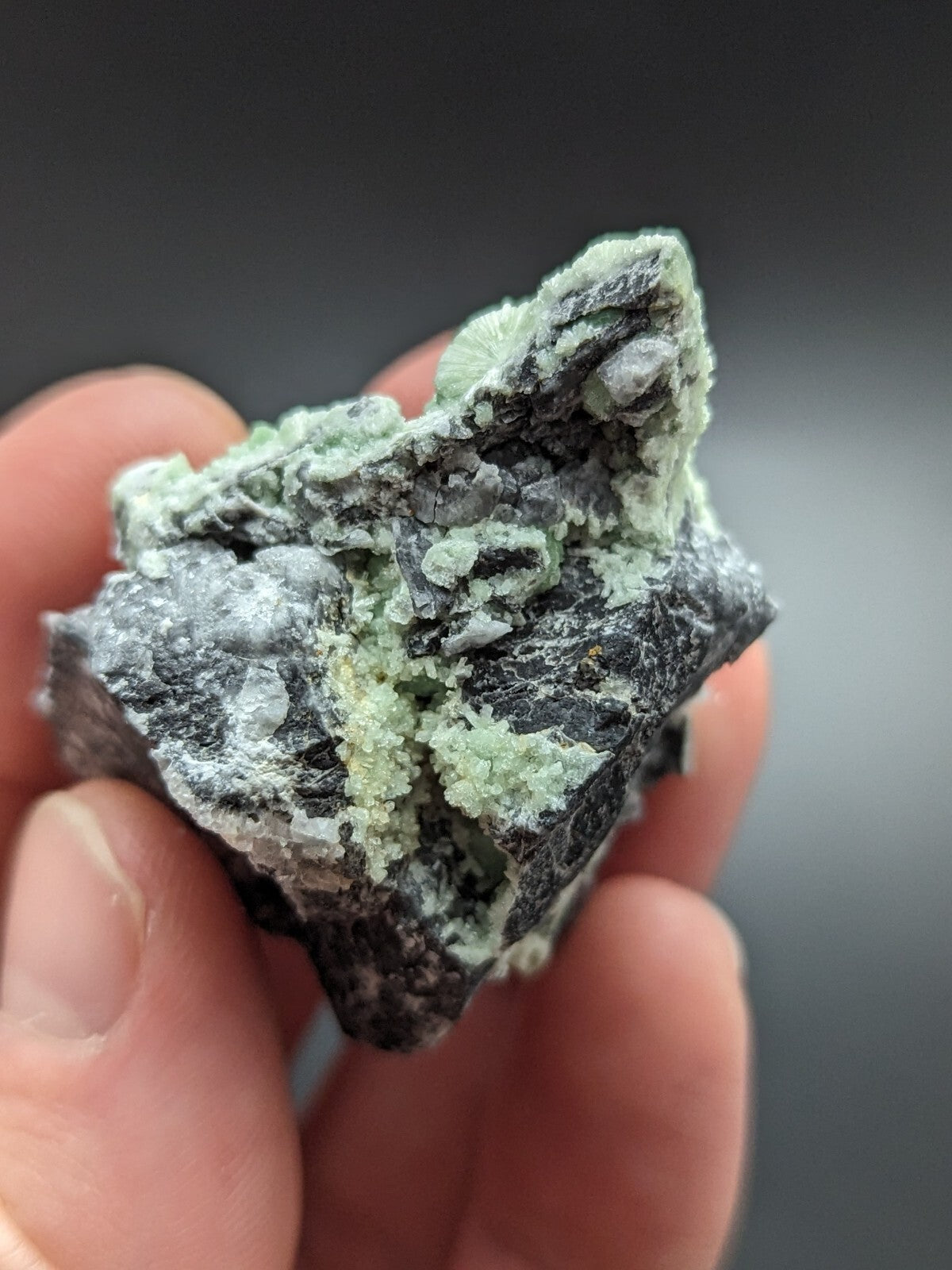A hand holds a rough, irregularly shaped mineral specimen featuring patches of pale green crystals growing over a dark gray matrix.