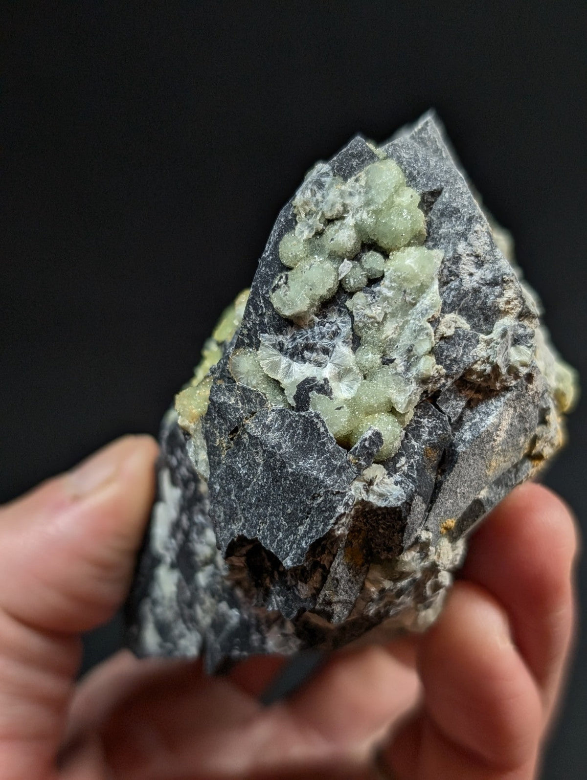 A hand holds a dark gray rock specimen featuring clusters of pale green, translucent crystals.