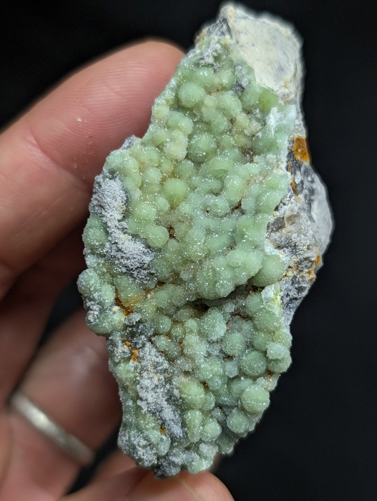 A hand holds a geode with clusters of translucent, light green crystals growing from a rough, grayish-white rock matrix.