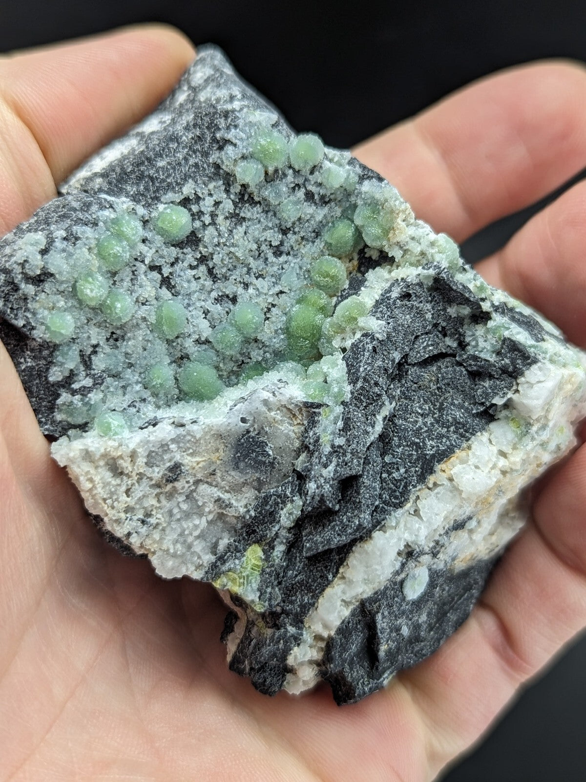 A hand holds a dark gray rock specimen featuring clusters of translucent, light green spherical crystals.