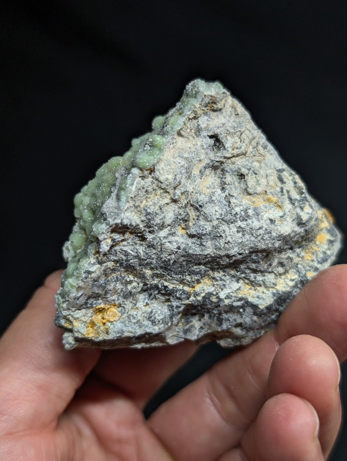 A hand holds a rough, irregularly shaped mineral specimen featuring patches of pale green crystals growing from a grayish matrix with subtle yellow flecks.