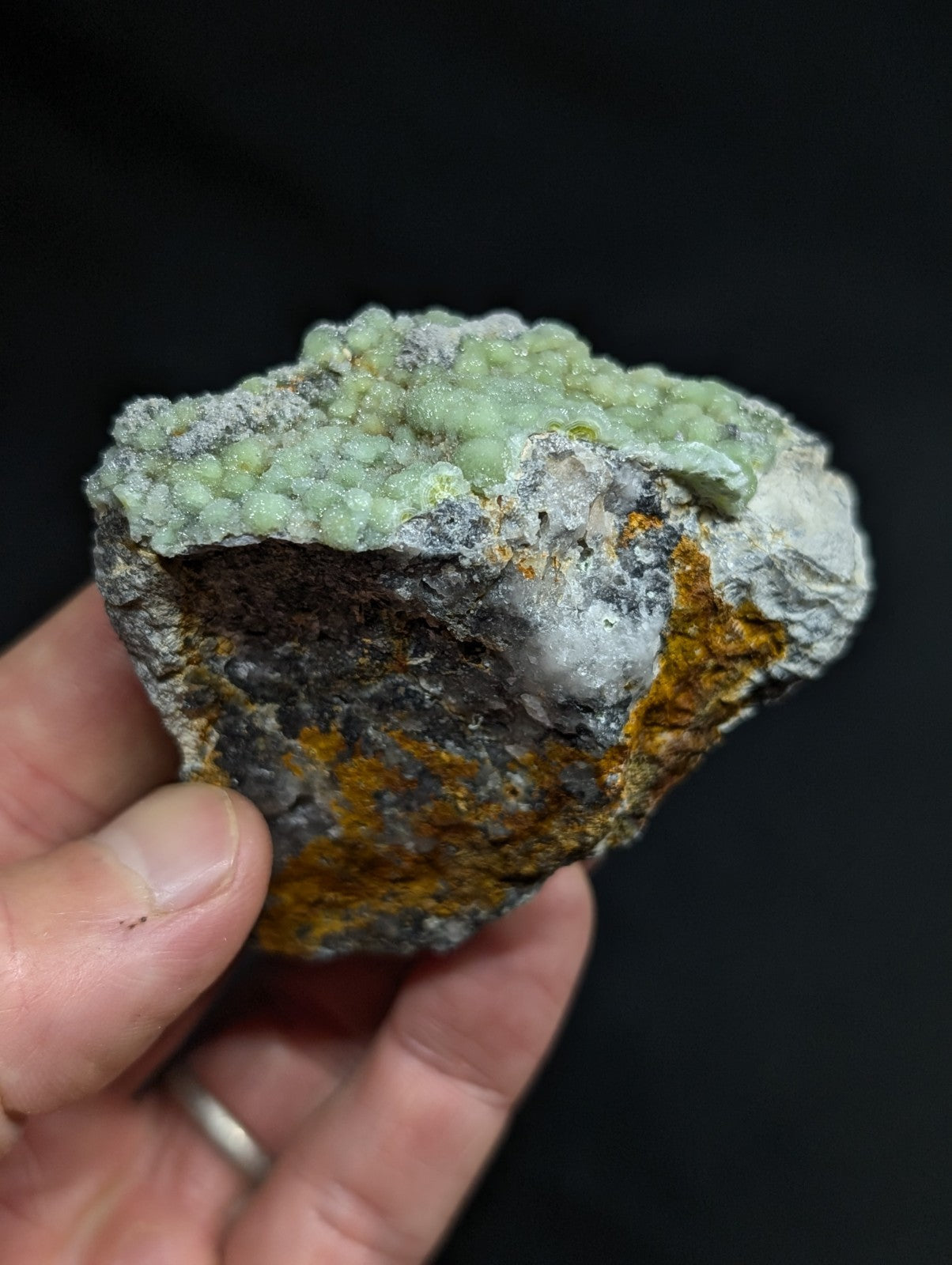 A hand holds a rough, irregularly shaped mineral specimen featuring a textured surface with patches of pale green crystalline growths and areas of grayish-white rock interspersed with streaks of orange-yellow oxidation.