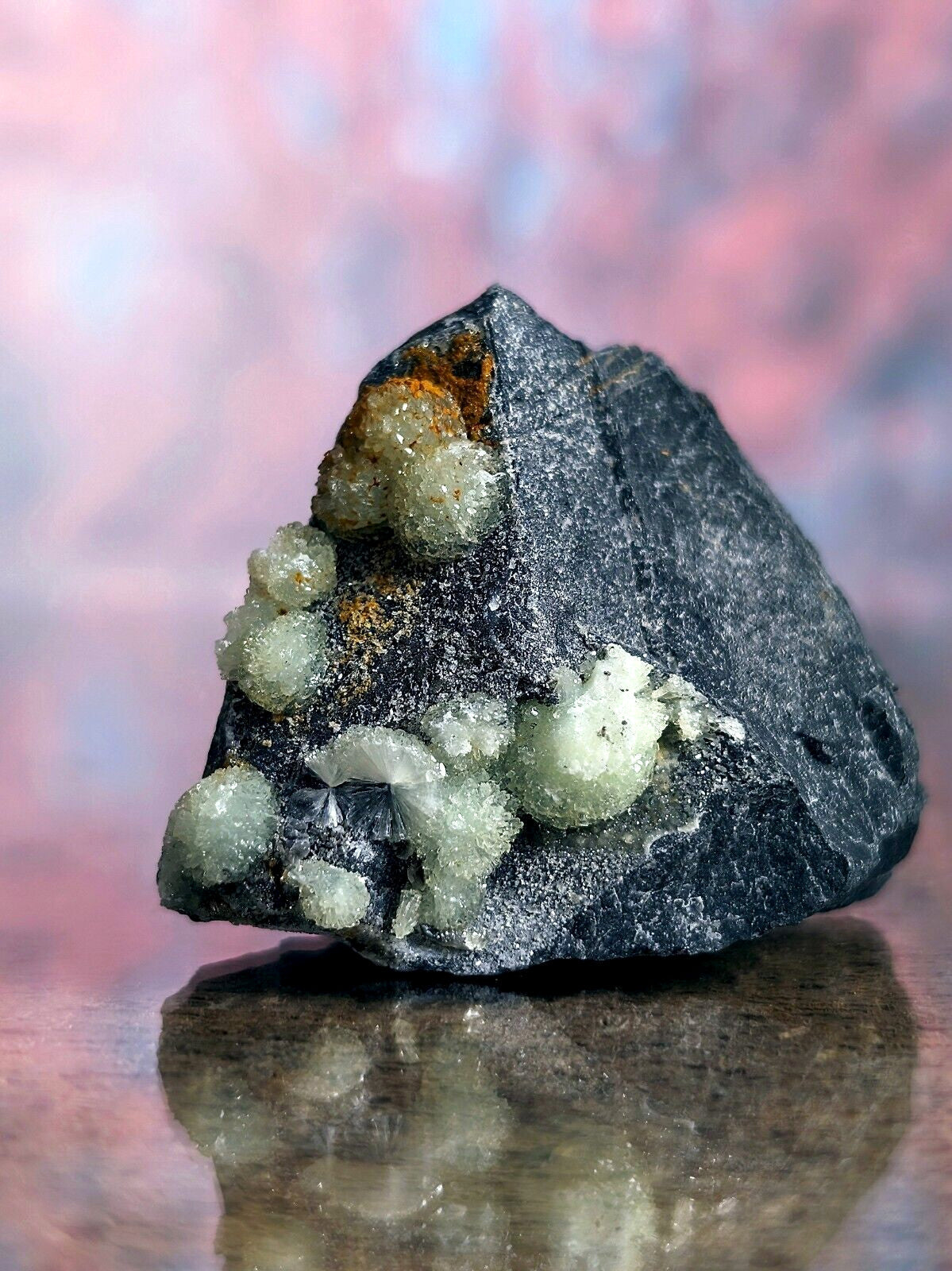 A dark gray rock specimen featuring clusters of translucent, light green crystalline formations with hints of orange mineral staining.