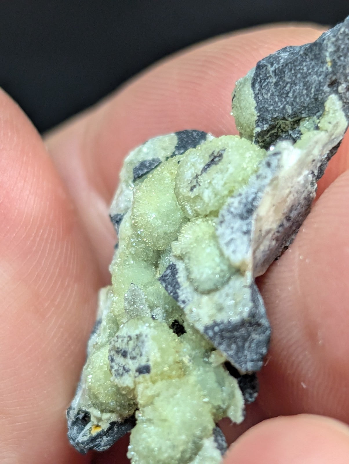 A hand holds a cluster of green and black mineral crystals with a rough, textured surface.