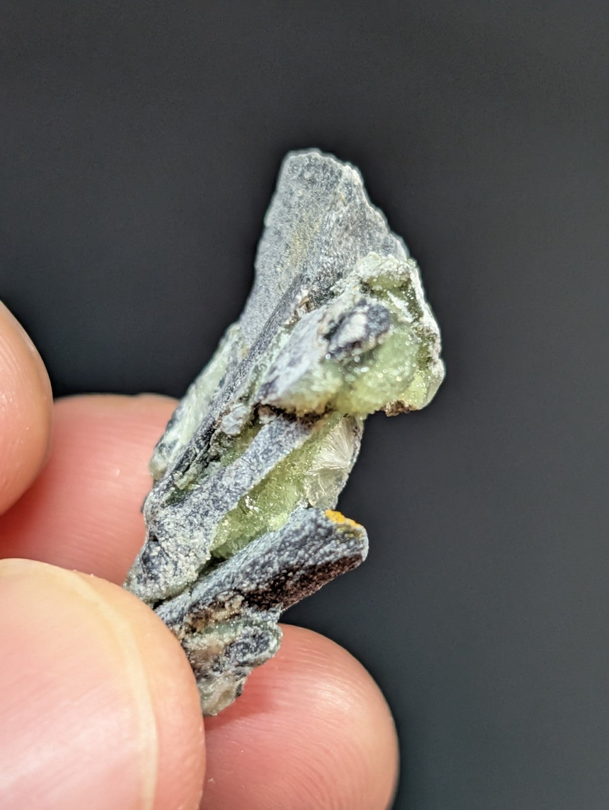 A rough, irregularly shaped green mineral specimen with a grayish matrix and visible crystalline facets.