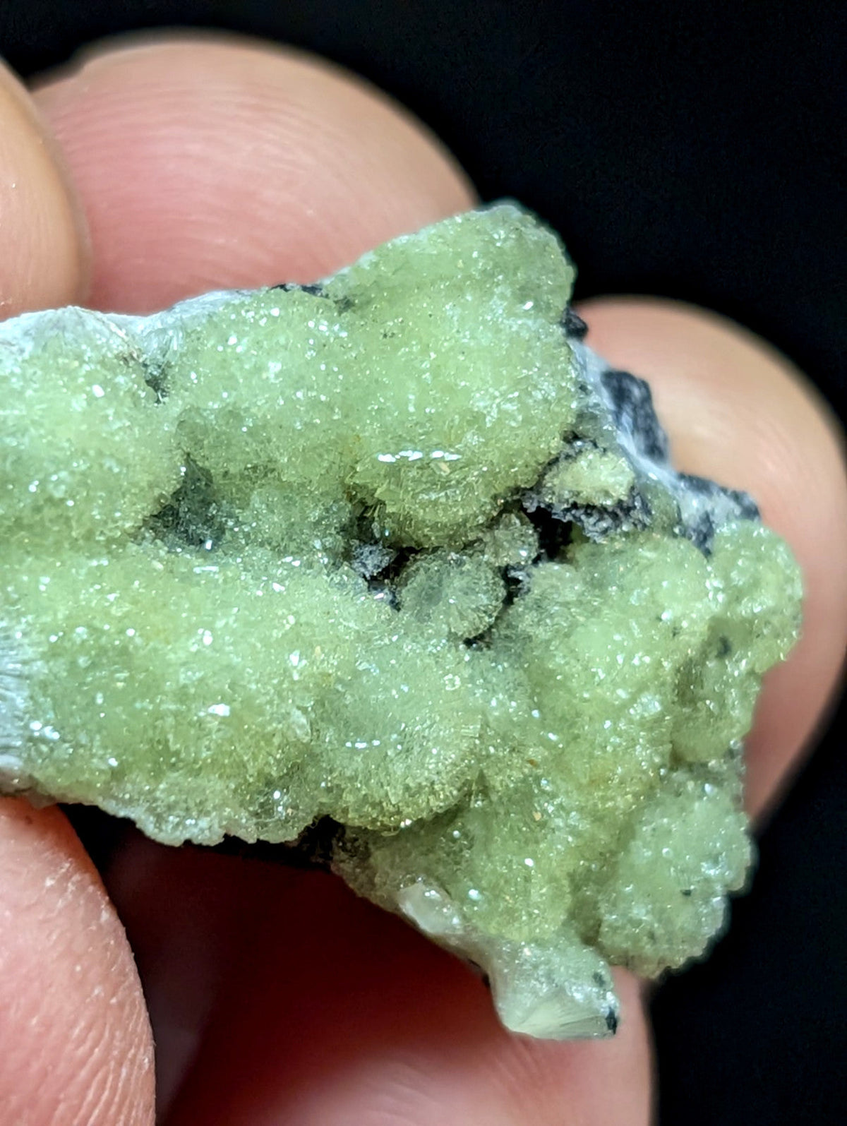 A pale green, crystalline mineral specimen with a rough, textured surface and embedded darker matrix.