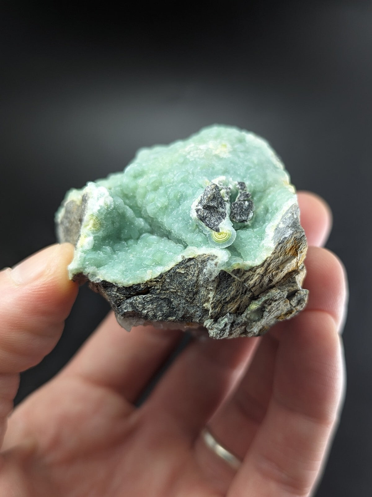 A hand holds a rough, irregularly shaped mineral specimen featuring a vibrant light green crystalline formation atop a dark gray rock base.