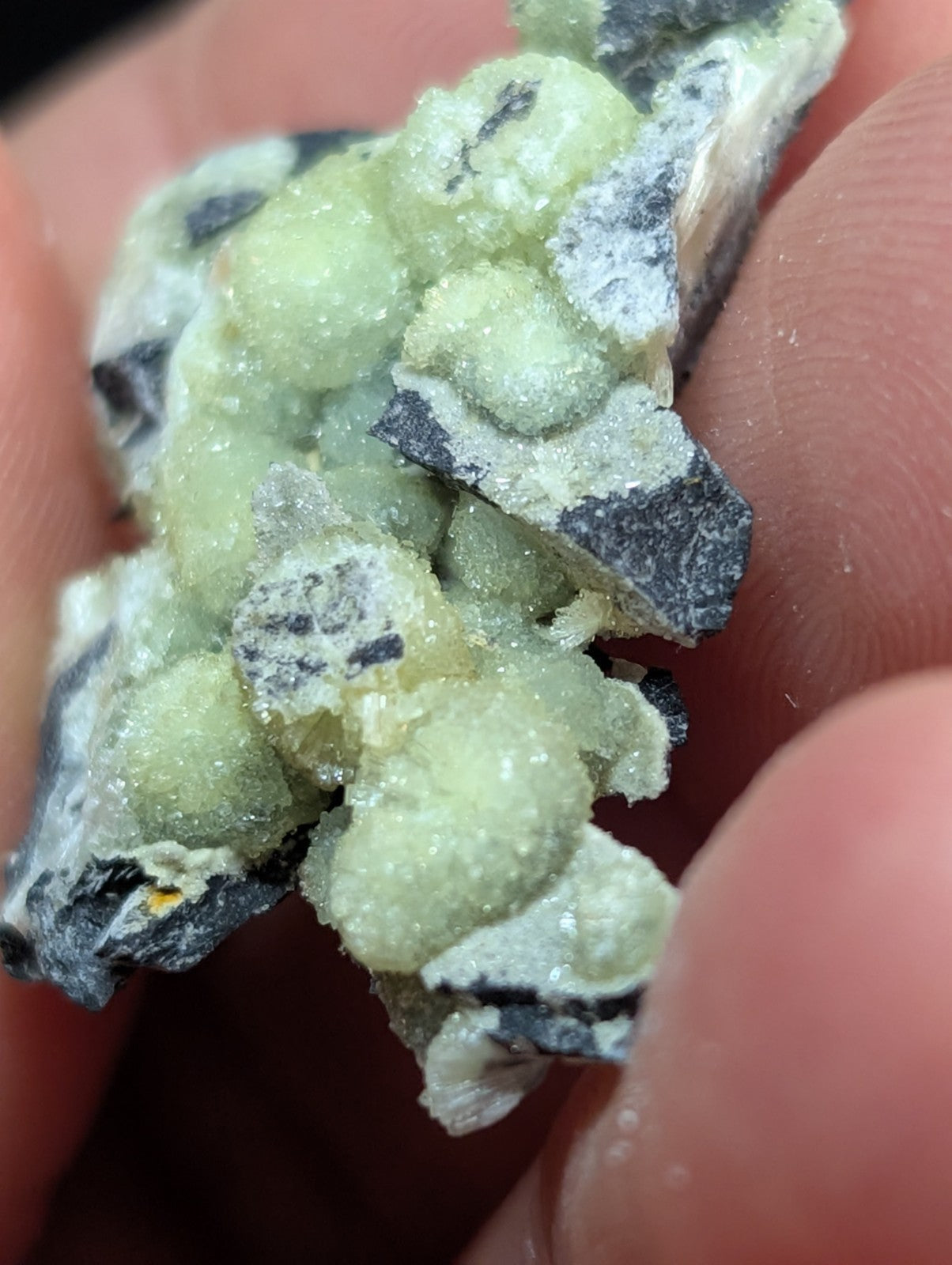 A cluster of pale green, crystalline mineral formations nestled within dark gray rock fragments.