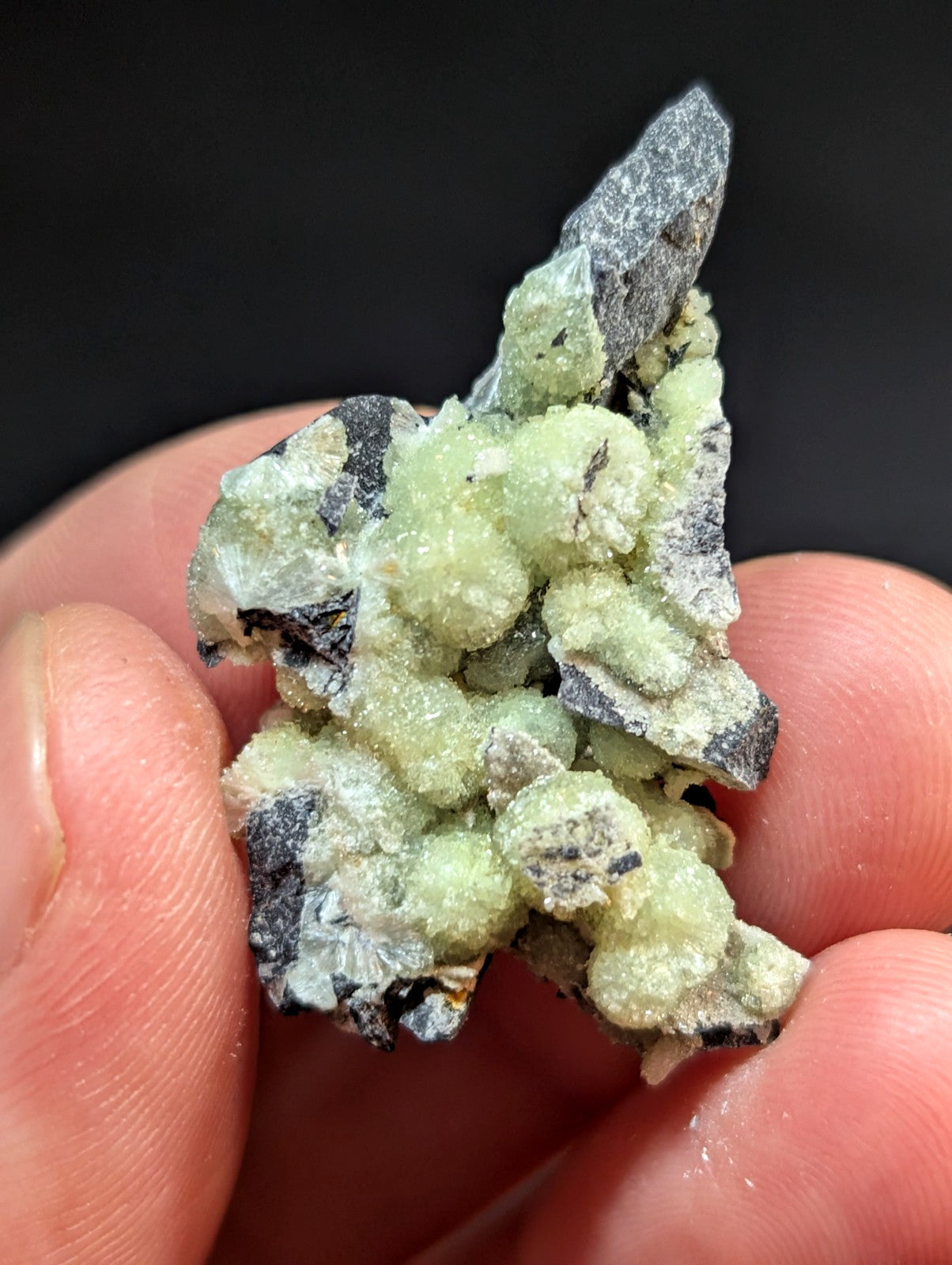 A cluster of pale green, crystalline minerals with a rough, textured surface and dark gray matrix.