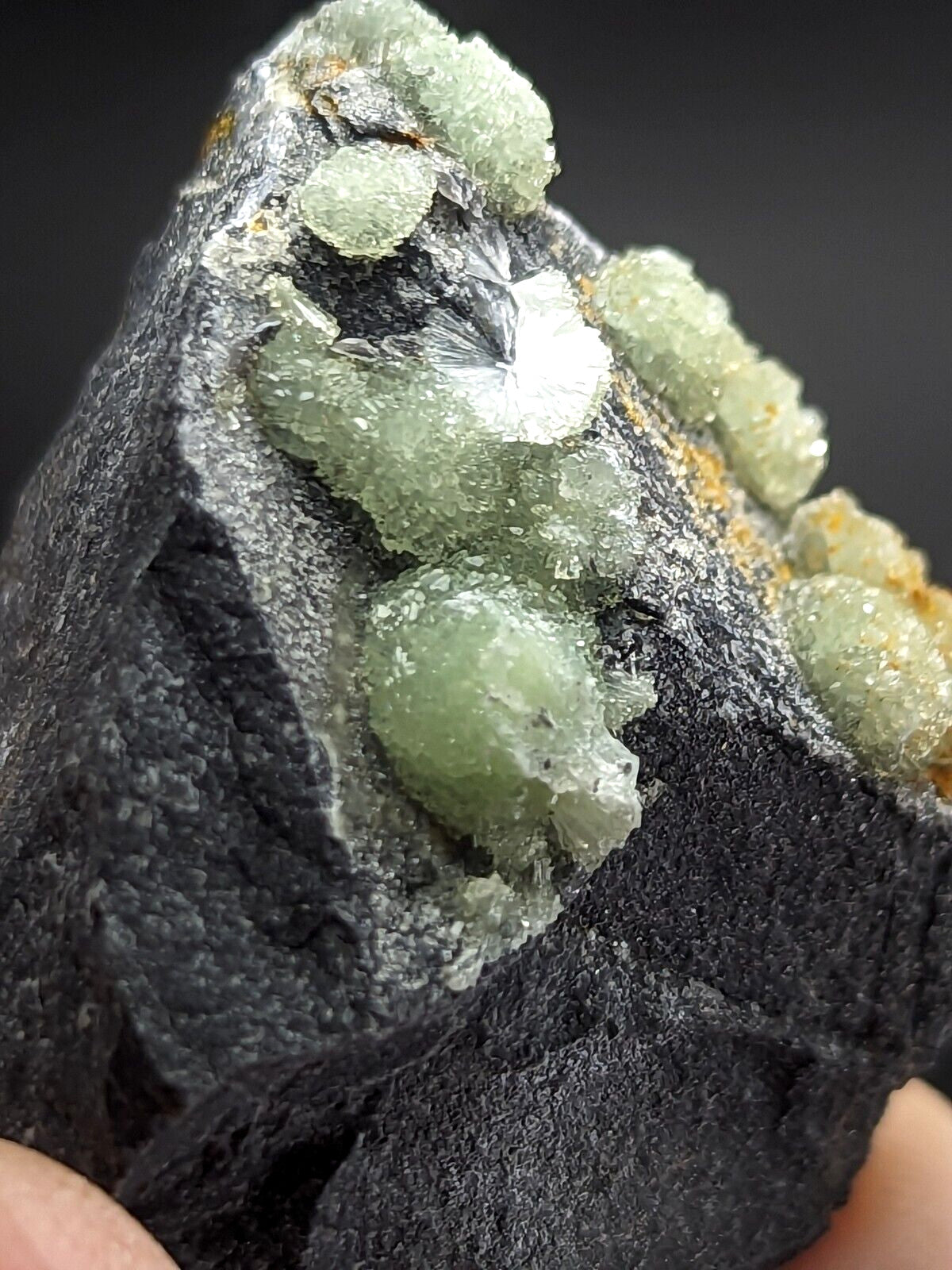 A dark, rough-textured rock host featuring clusters of translucent green crystals with a crystalline, faceted appearance.