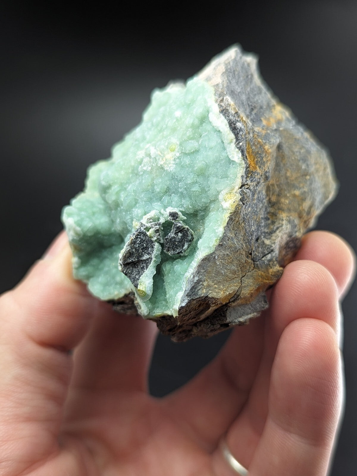 A hand holds a rough, irregularly shaped mineral specimen featuring vibrant light green crystalline formations against darker gray and brown rock matrix.