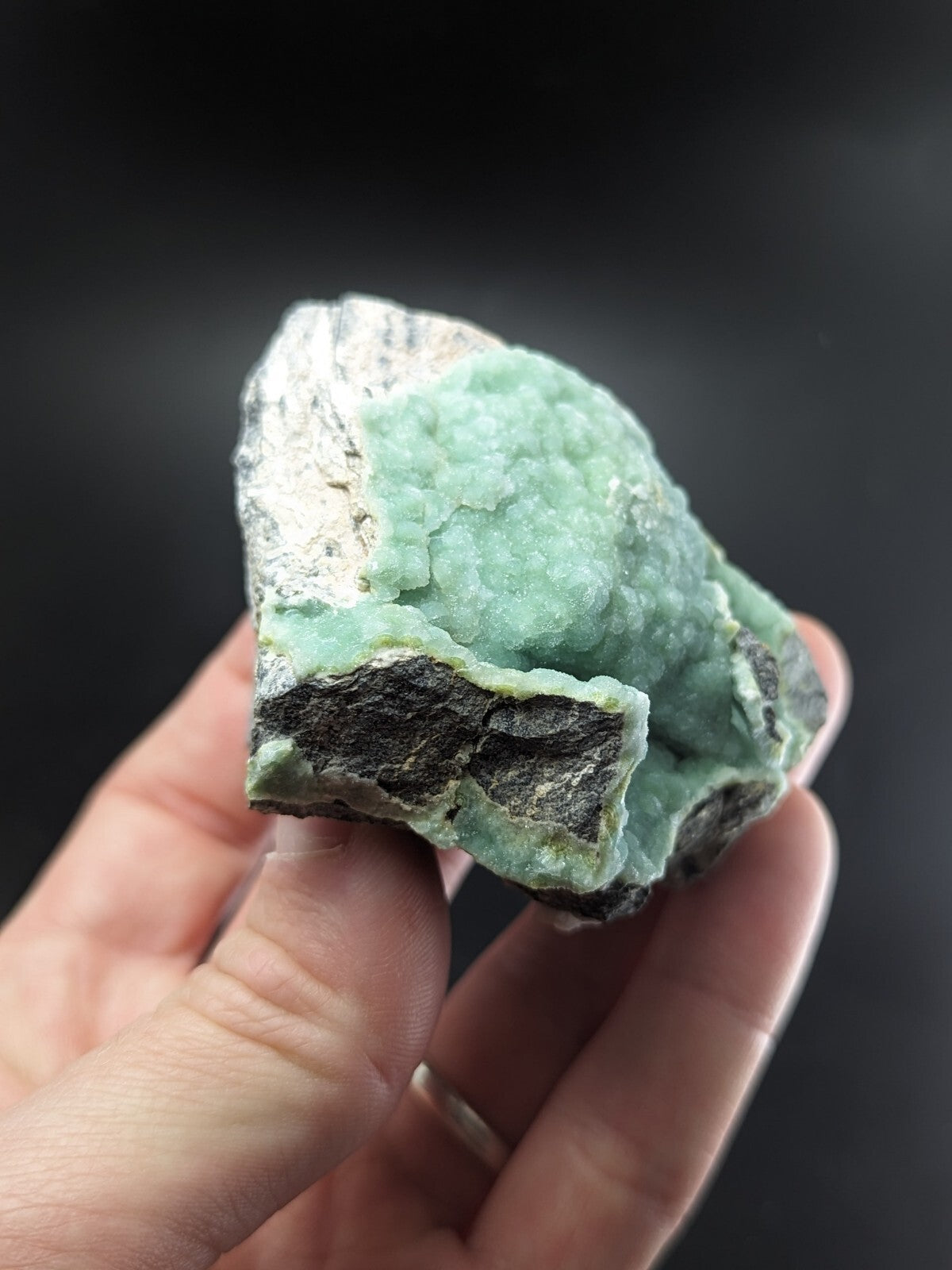 A hand holds a rough, irregularly shaped mineral specimen featuring vibrant light green crystalline formations overlying darker gray and white matrix material.