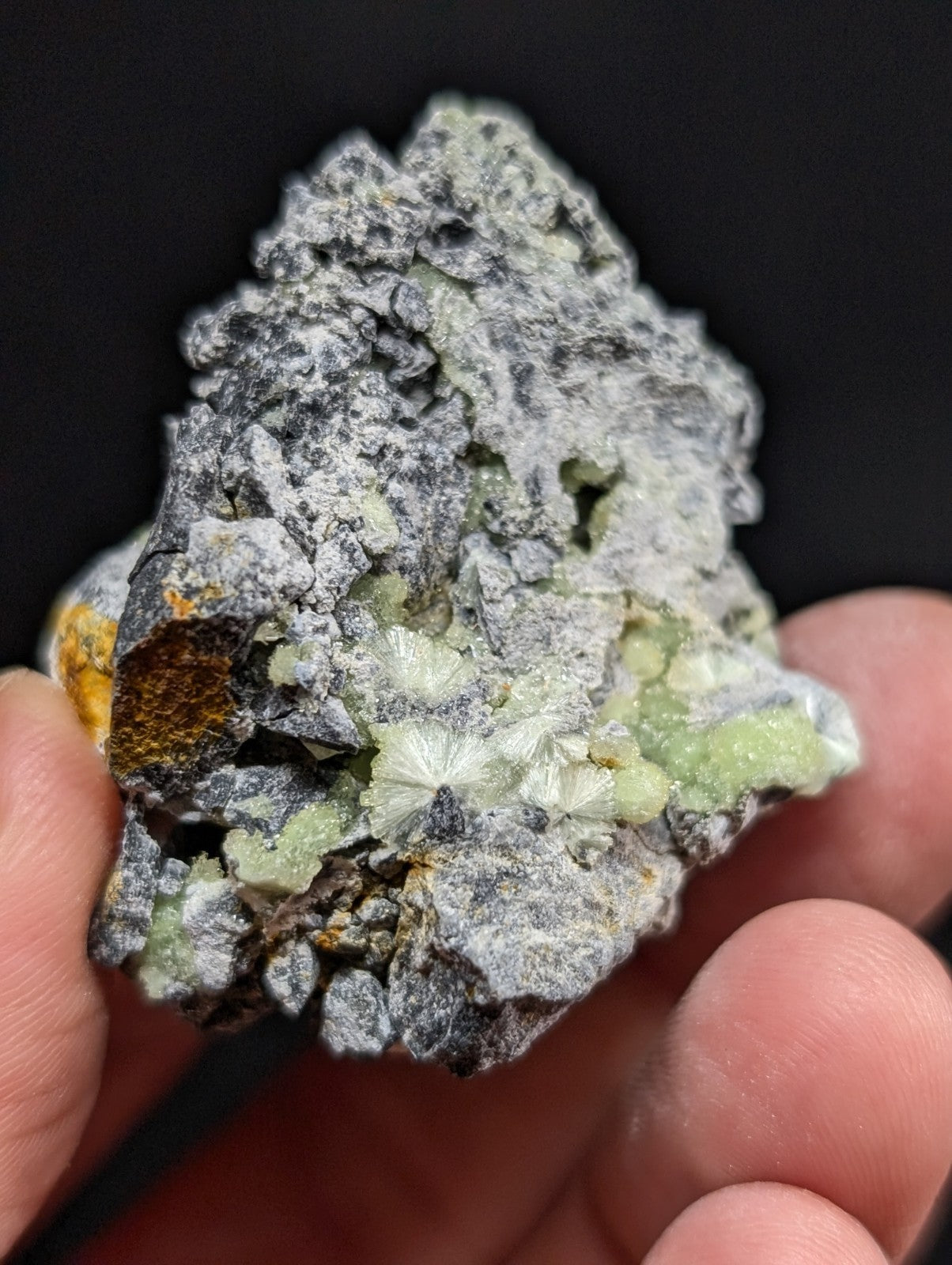 A hand holds a rough, grayish rock specimen featuring clusters of pale green, crystalline formations and patches of orange mineral.