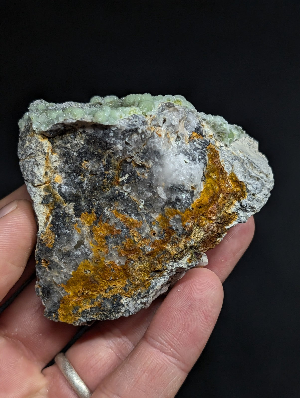 A hand holds a rough, irregularly shaped mineral specimen featuring patches of gray rock with vibrant orange and pale green crystalline formations.