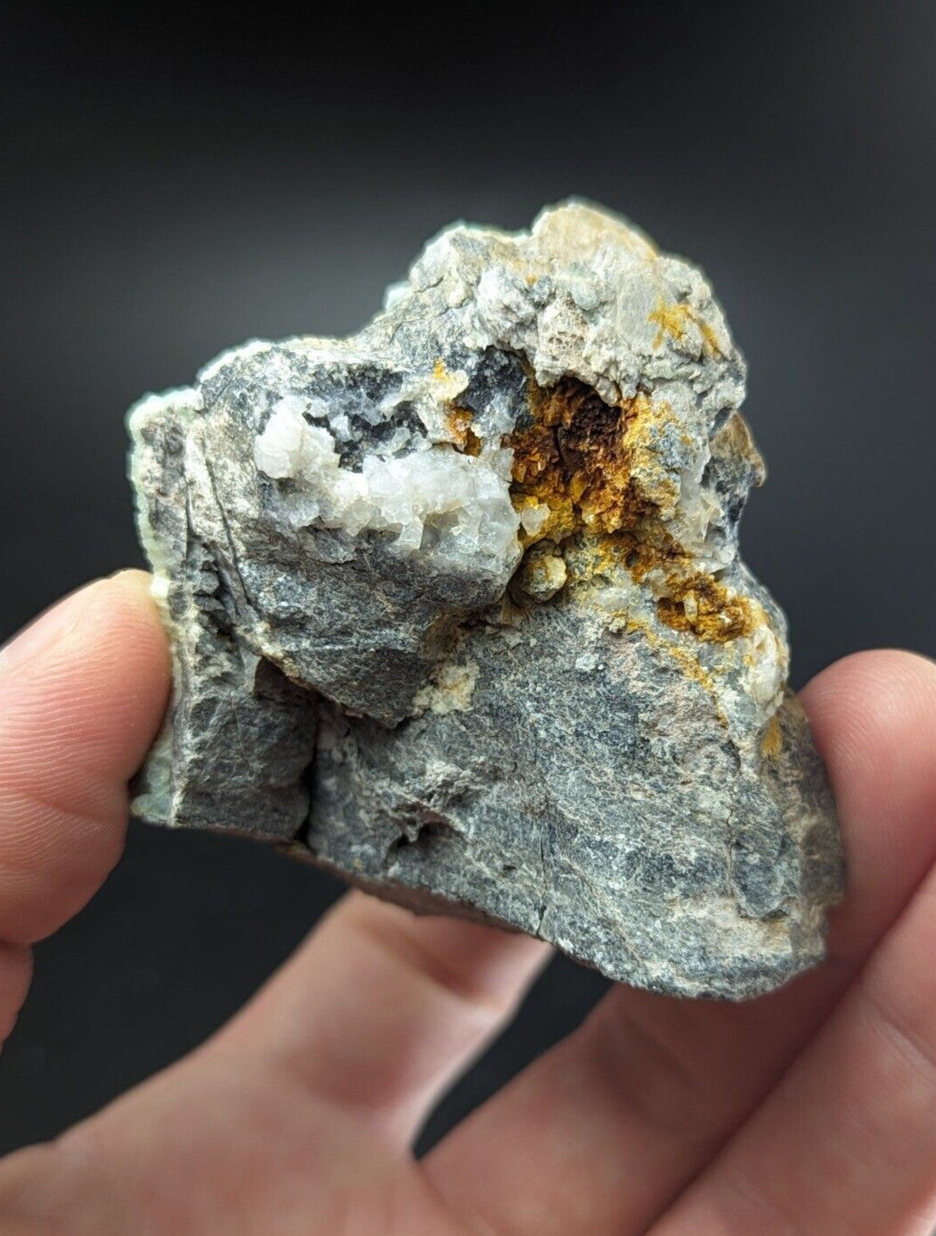 A rugged gray rock specimen with a textured surface, featuring patches of white crystalline formations and vibrant orange-yellow mineral inclusions.