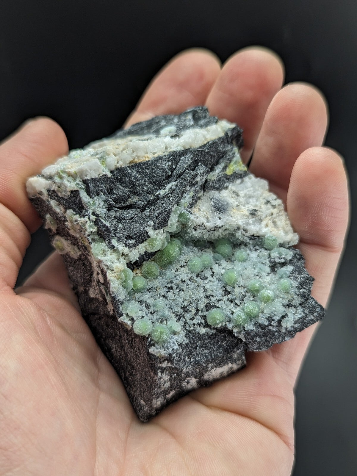 A hand holds a dark, rough-textured rock specimen featuring clusters of small, translucent green crystals embedded in its surface.