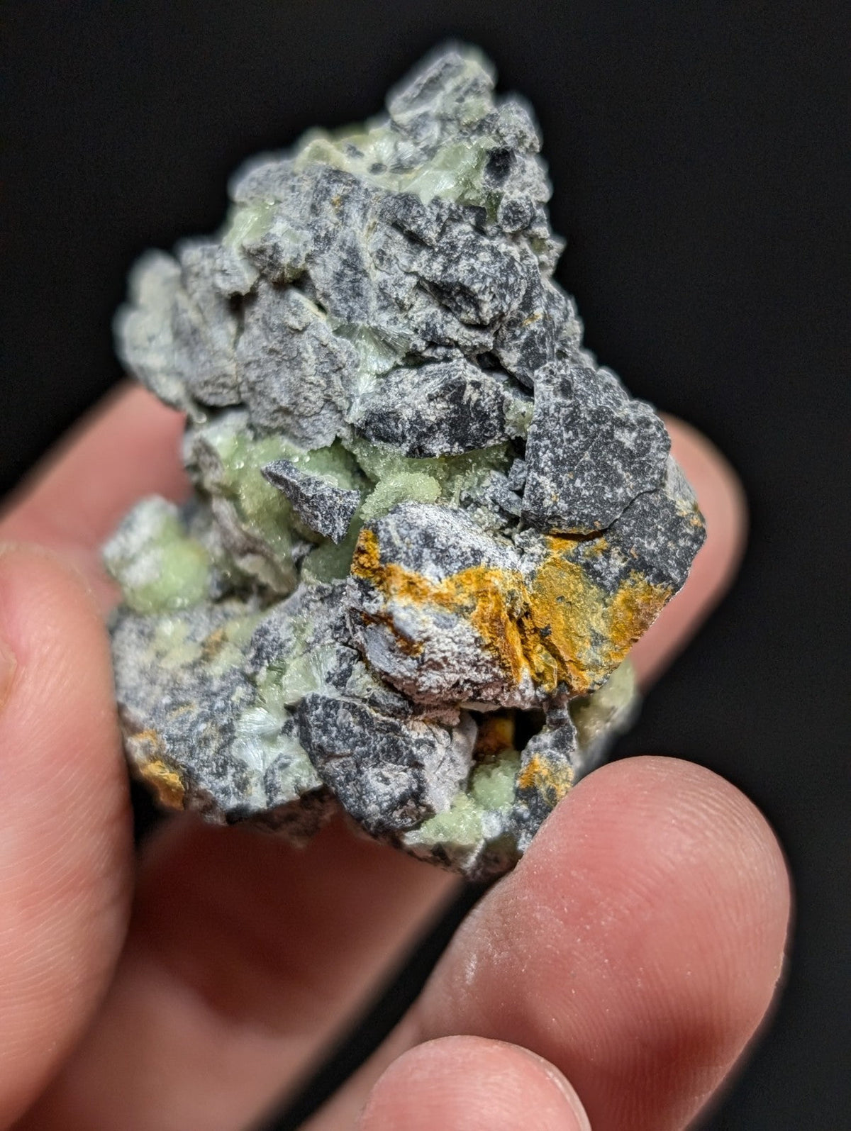 A rough, irregularly shaped mineral specimen featuring a mix of gray and pale green crystalline formations with patches of vibrant orange-yellow streaks.