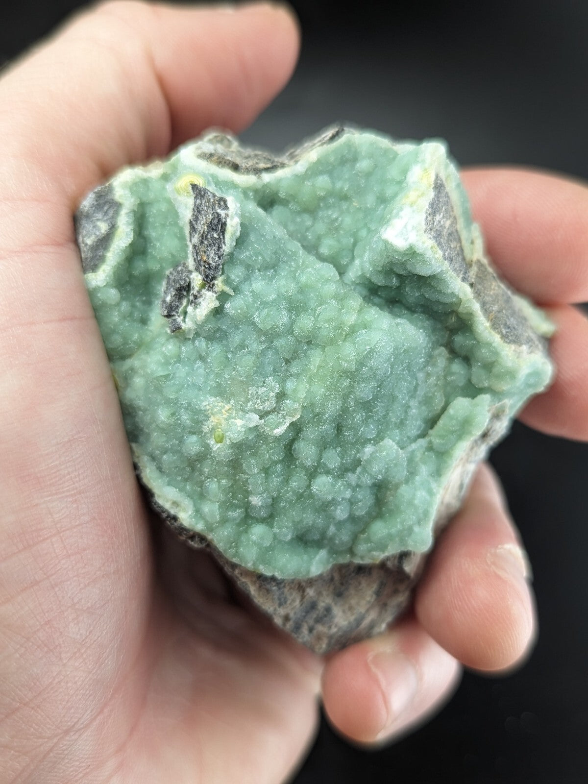 A hand holds a rough, light green mineral specimen with small crystalline formations and dark gray patches.