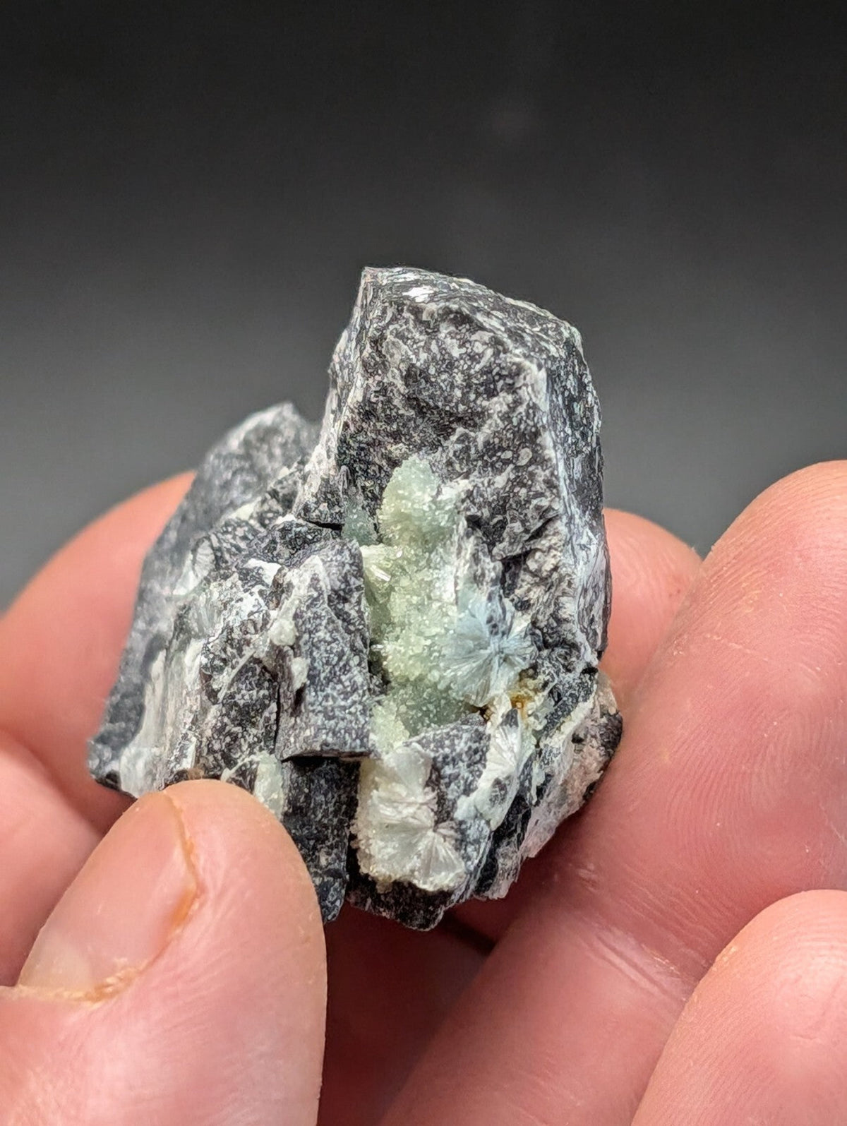 A hand holds a rough, dark gray rock with embedded translucent light green crystals.