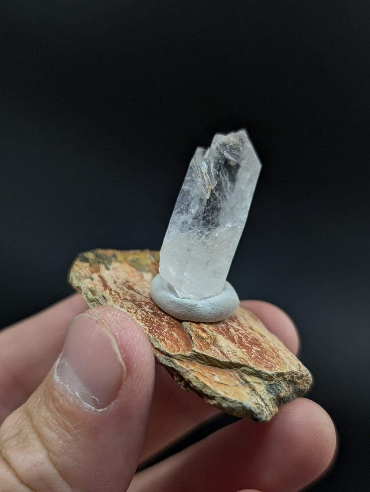 A clear quartz crystal mounted on a natural rock base with a light gray circular holder.