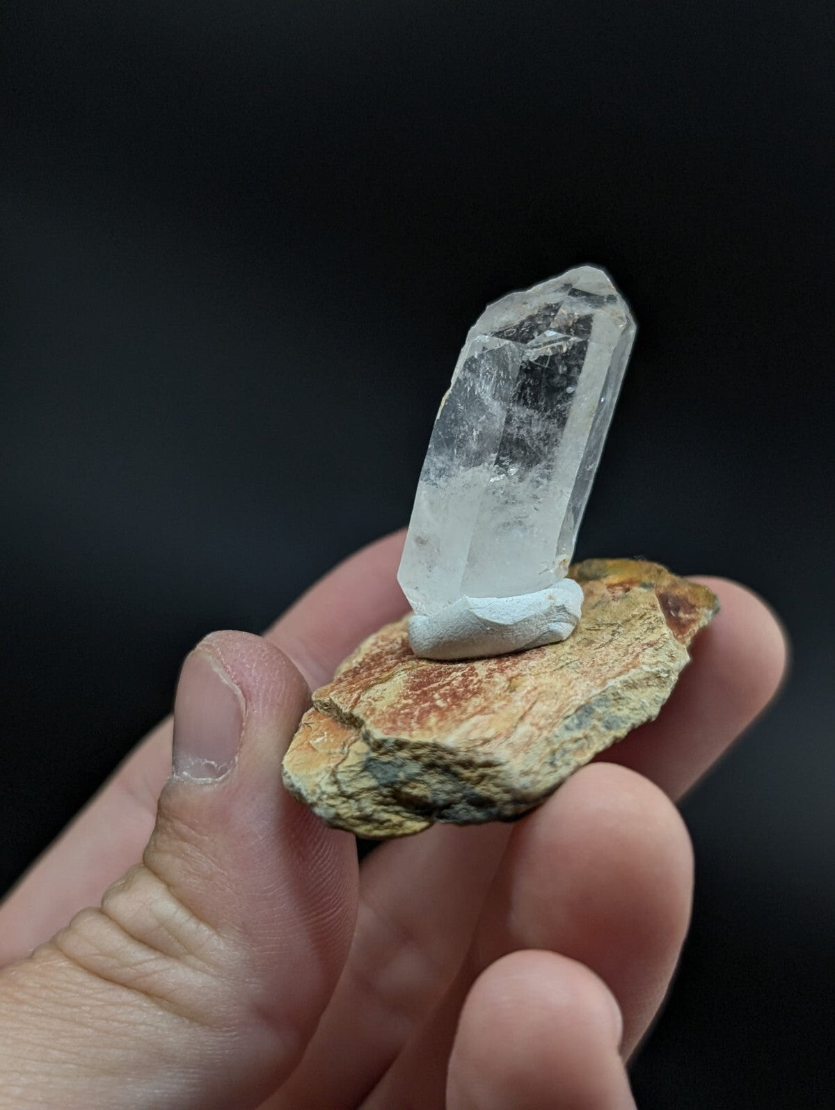 A clear quartz crystal with a pointed tip, mounted on a textured brown and yellow rock base.