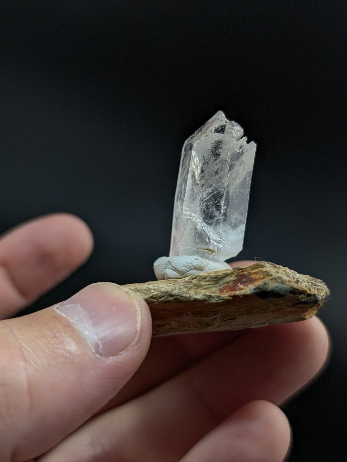 A clear, translucent quartz crystal with a pointed tip and visible internal inclusions is mounted on a rough, earthy-toned rock base.