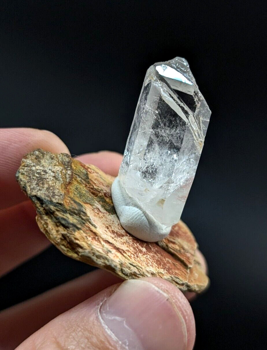 A clear, translucent quartz crystal with a pointed tip and visible internal inclusions is held upright on a rough-textured brownish rock.