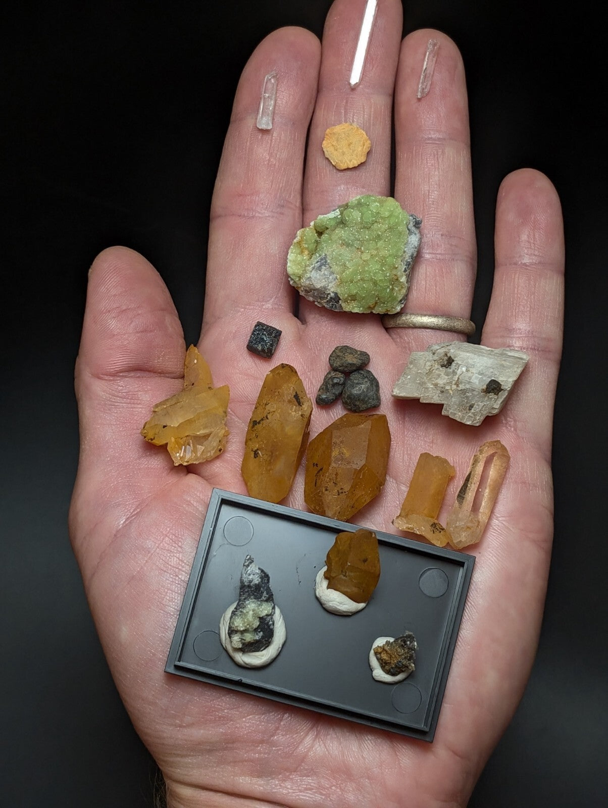 A hand holds a collection of colorful, naturally occurring minerals and crystals in various shapes, sizes, and hues including green, orange, yellow, gray, and clear.