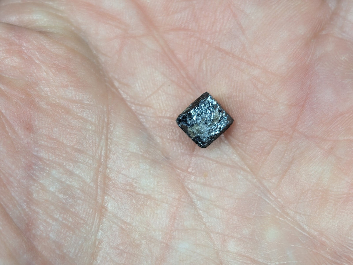 A small, dark metallic cube with a rough, textured surface rests in the palm of a hand.