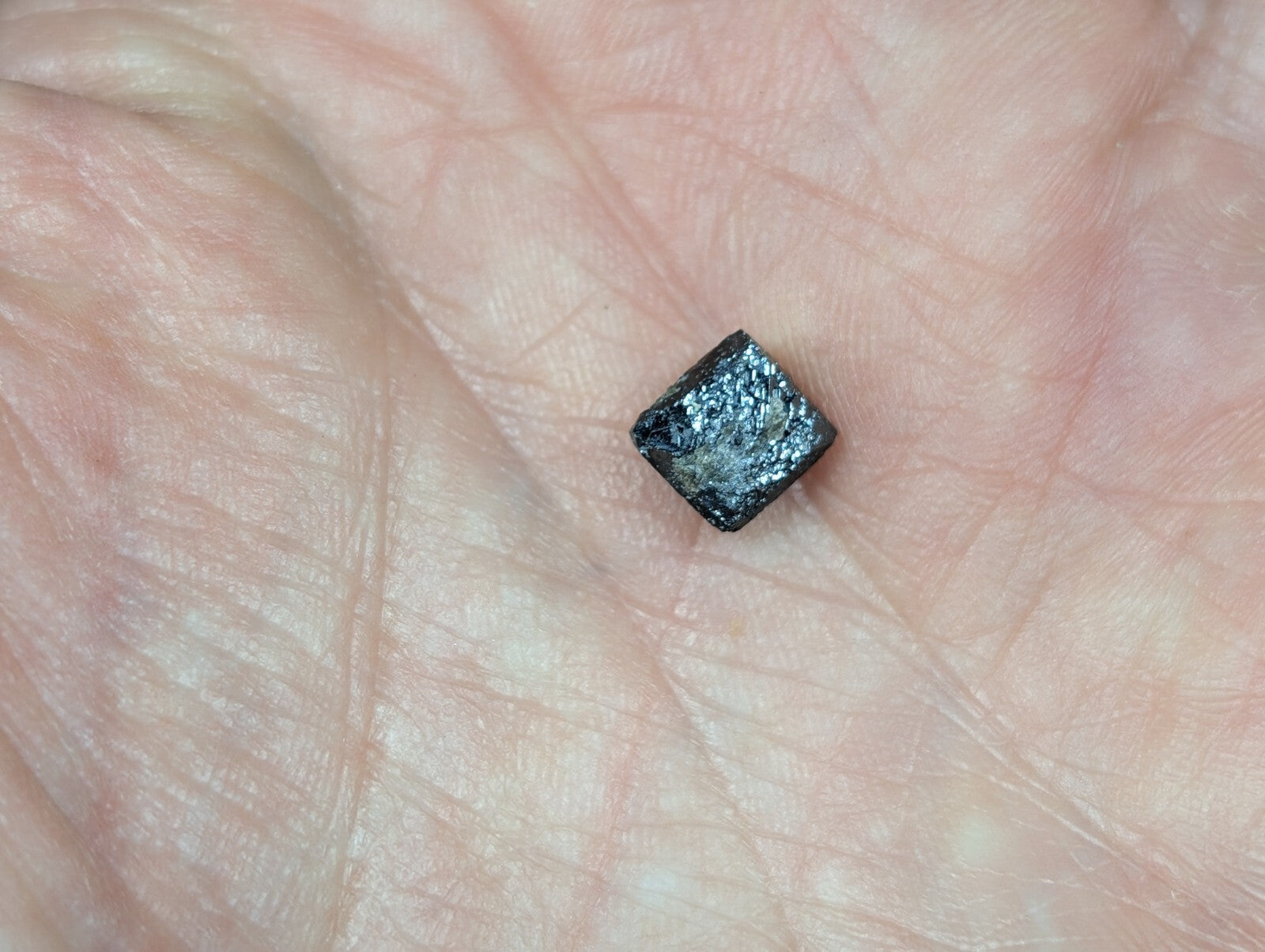 A small, dark metallic cube with a rough, textured surface rests in the palm of a hand.