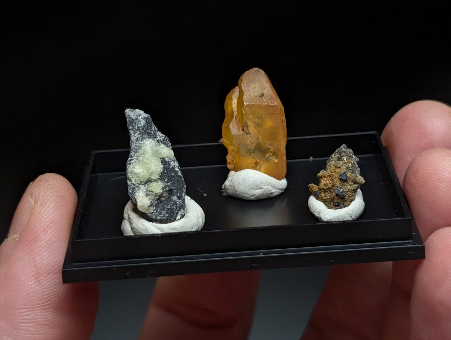 A black display tray holding three distinct mineral specimens, each mounted on a white base.