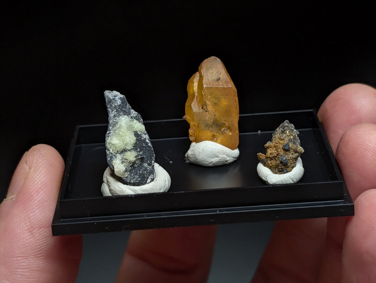 A black display tray holding three distinct mineral specimens, each mounted on a white base.