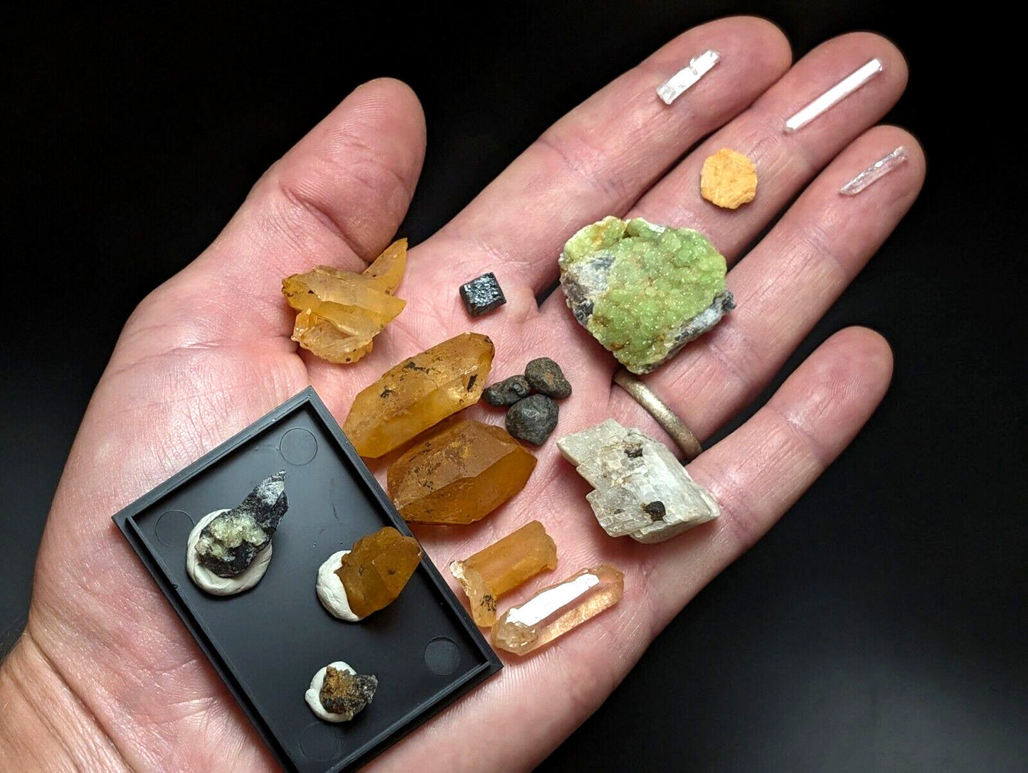 A hand holds a collection of colorful, naturally formed mineral specimens including amber-colored crystals and green crystalline formations alongside small white rectangular pieces.