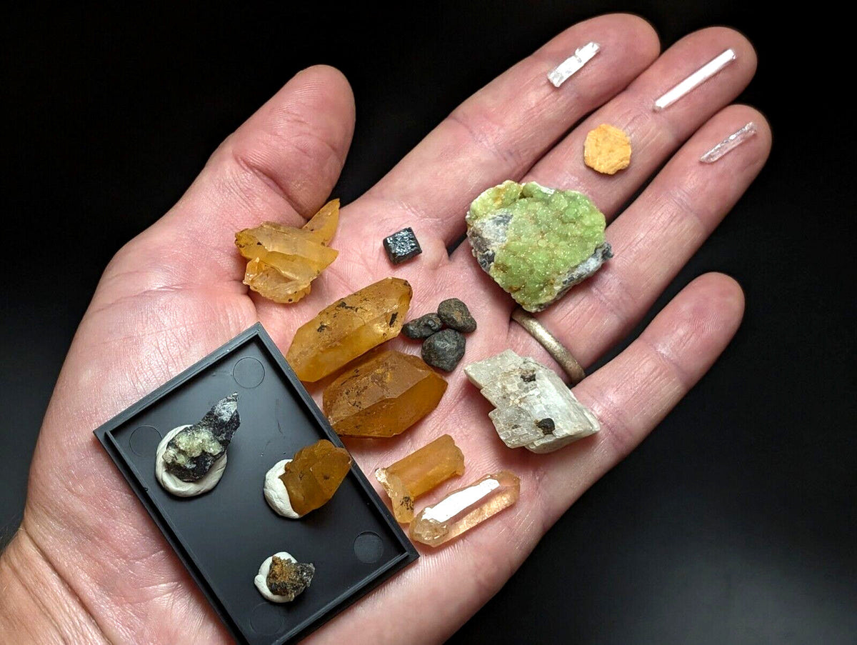 A hand holds a collection of colorful, naturally formed mineral specimens including amber-colored crystals and green crystalline formations alongside small white rectangular pieces.