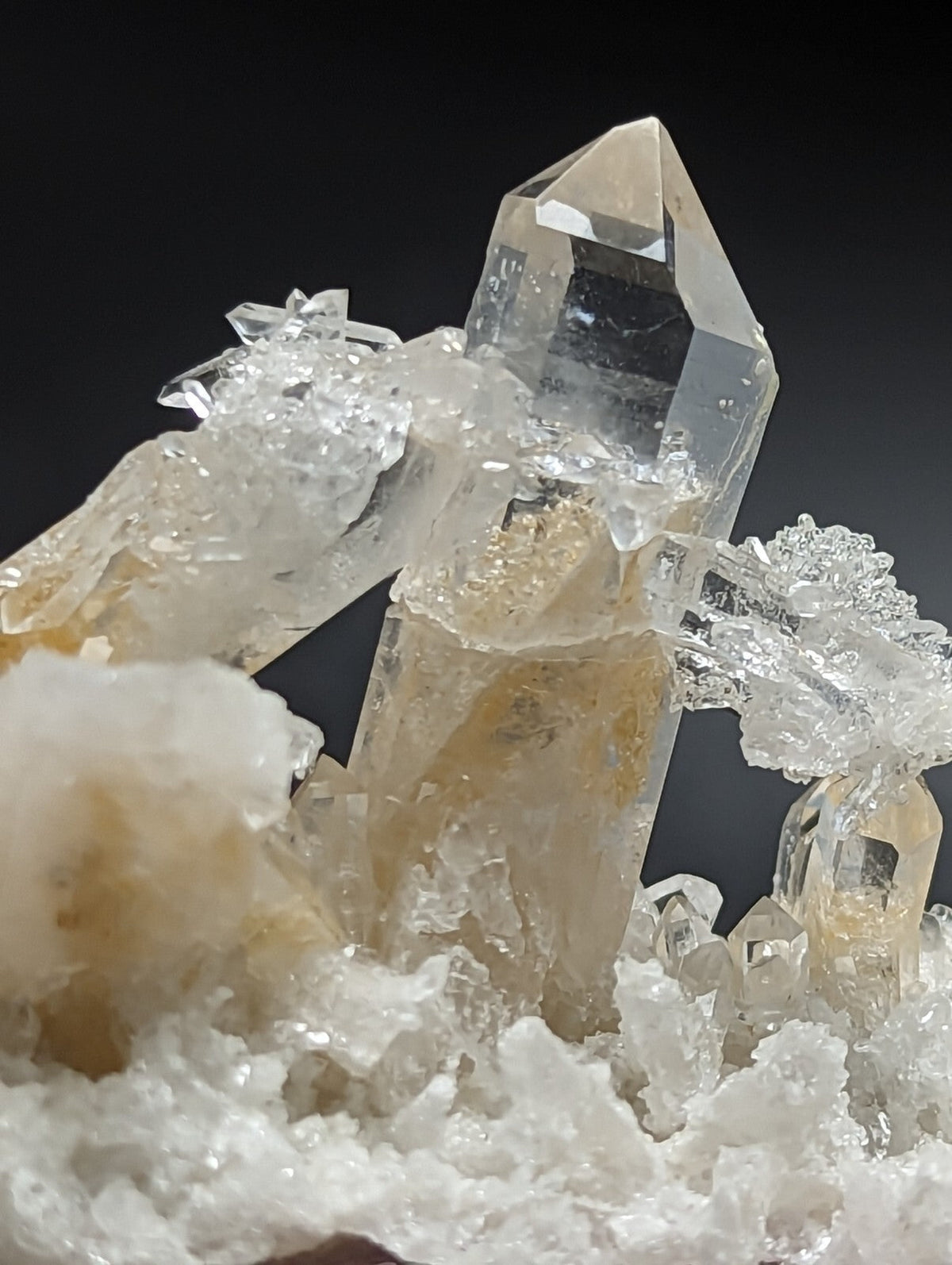 A clear quartz crystal cluster with a prominent tall, faceted point and surrounding smaller crystals embedded in a bed of white matrix material.