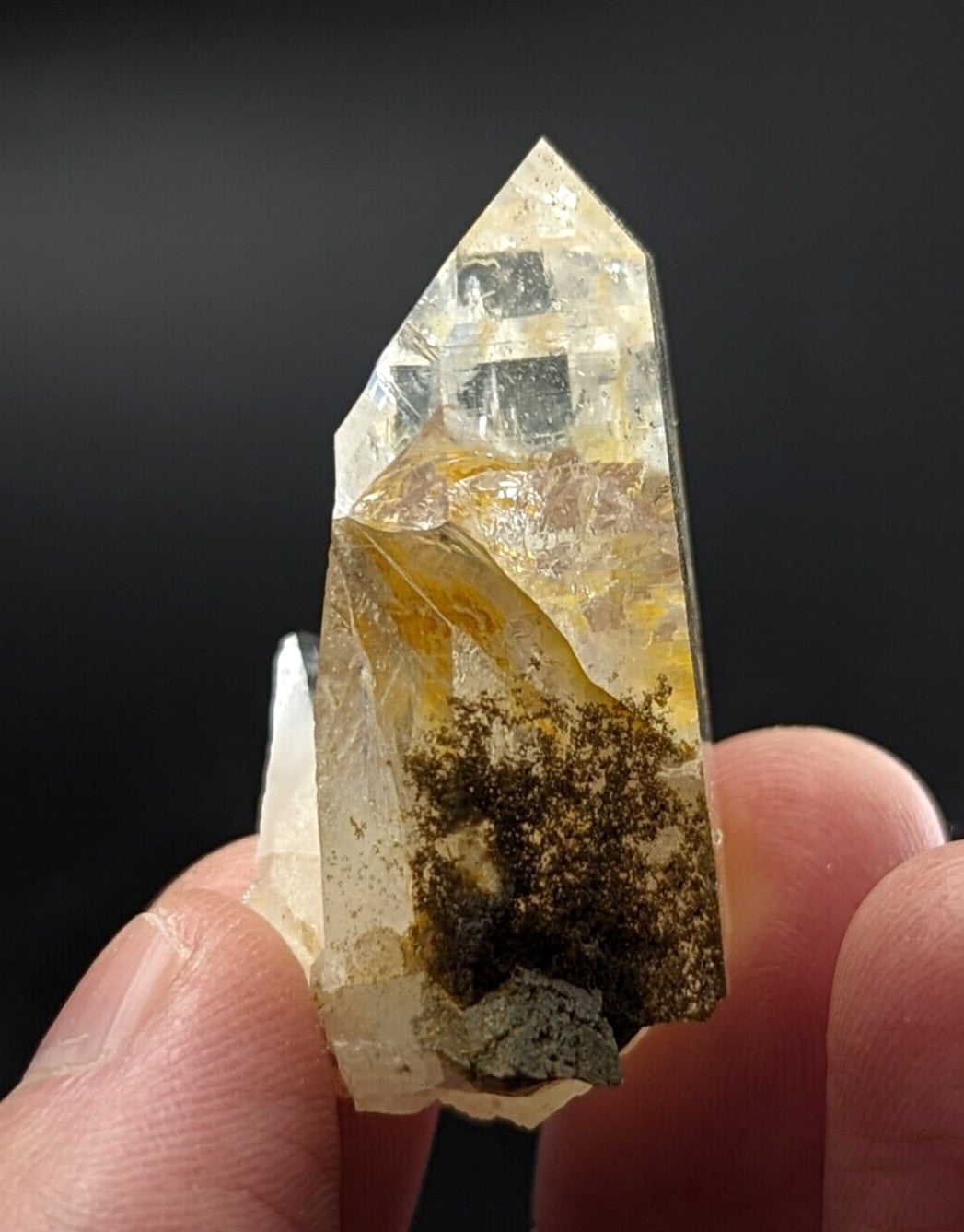A translucent yellowish quartz crystal with a pointed tip and internal brown inclusions, held between fingers.