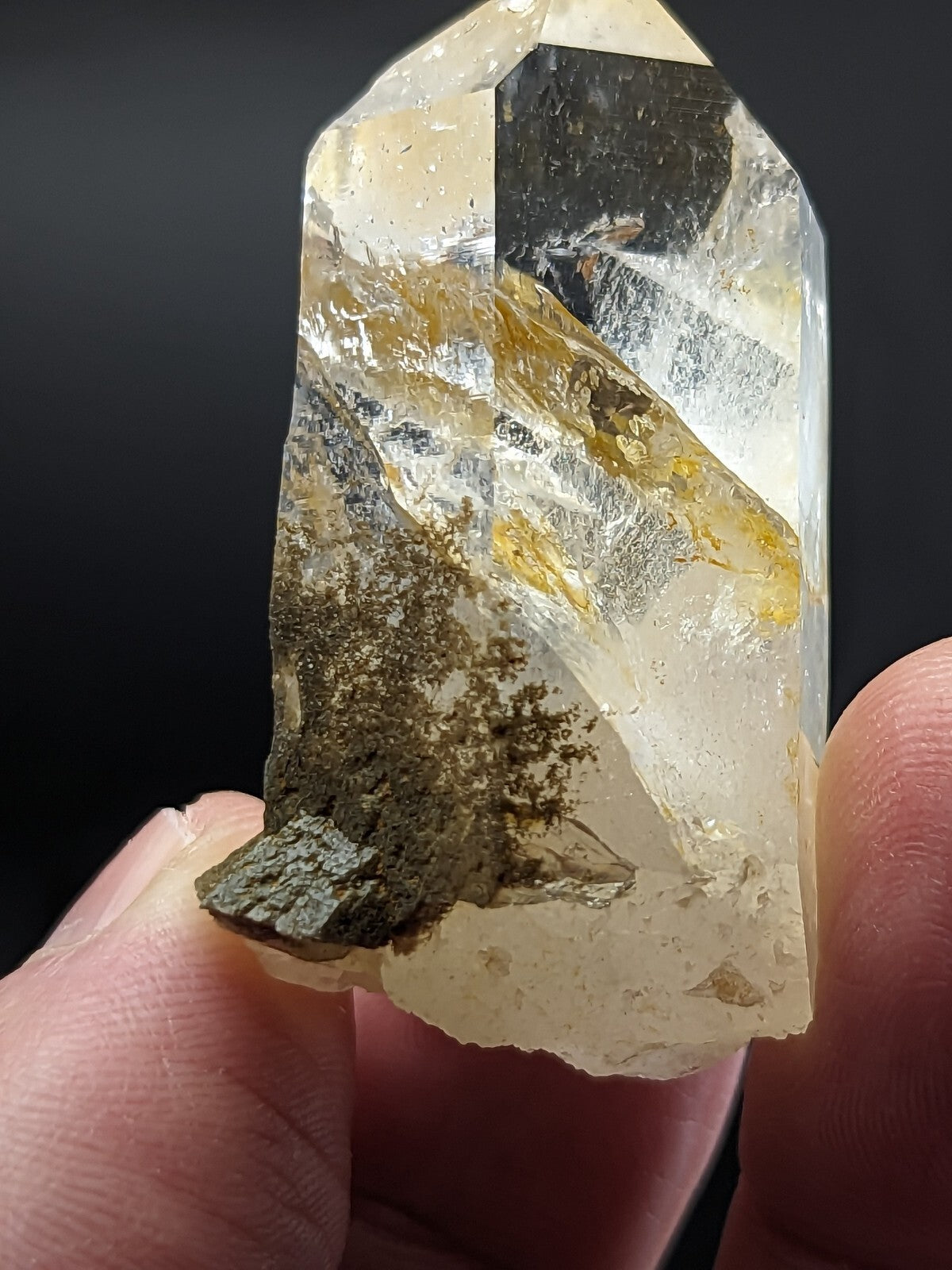 A translucent, pale yellow quartz crystal with golden inclusions and a dark mineral base held gently between fingers.