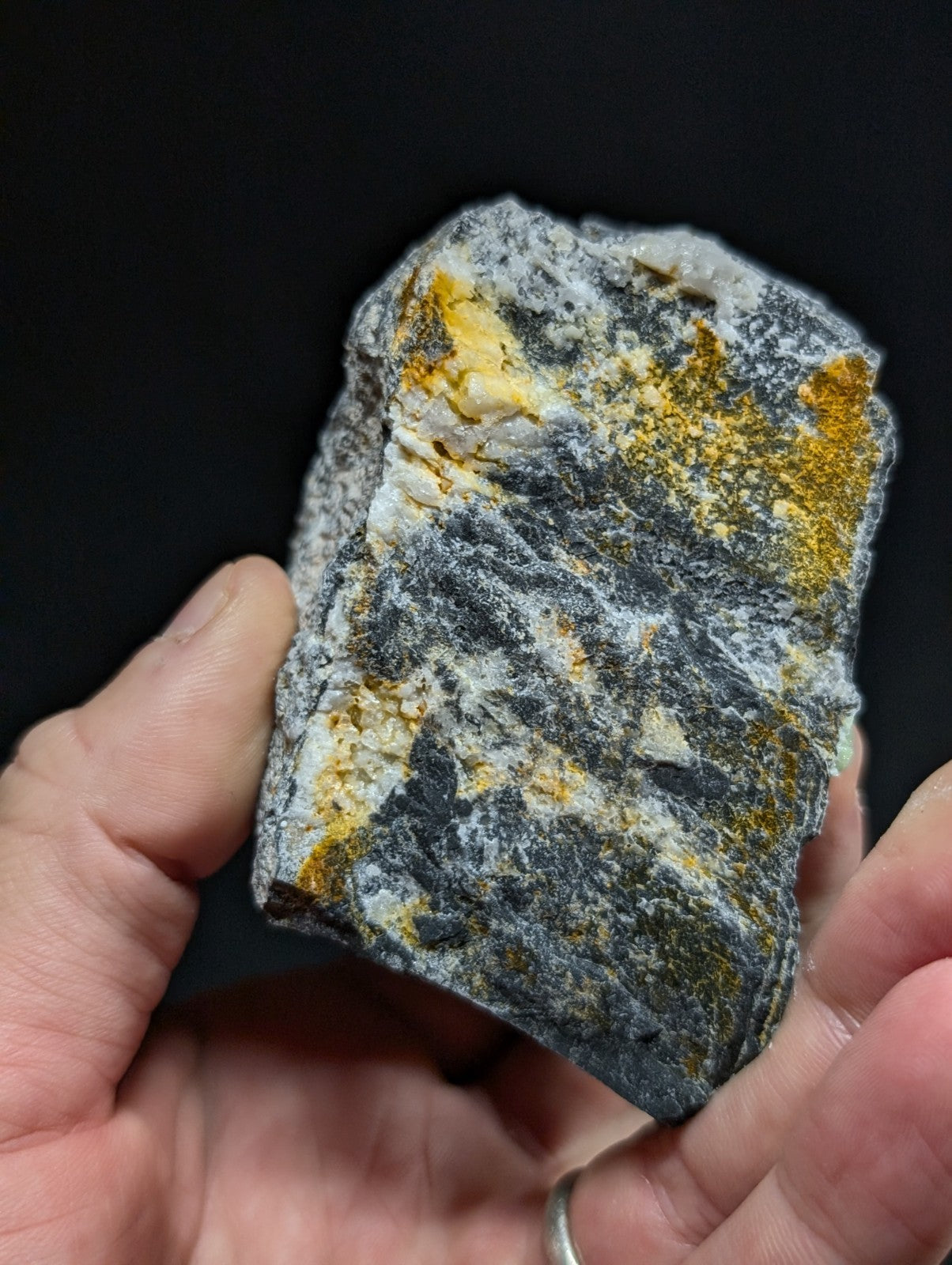 A hand holds a rough, irregularly shaped mineral specimen with dark gray and white matrix veined by patches of vibrant yellow-orange oxidation.