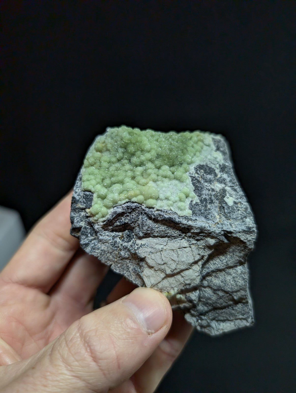 A hand holds a geological specimen featuring a textured, grayish rock base with a vibrant green crystalline formation on top.