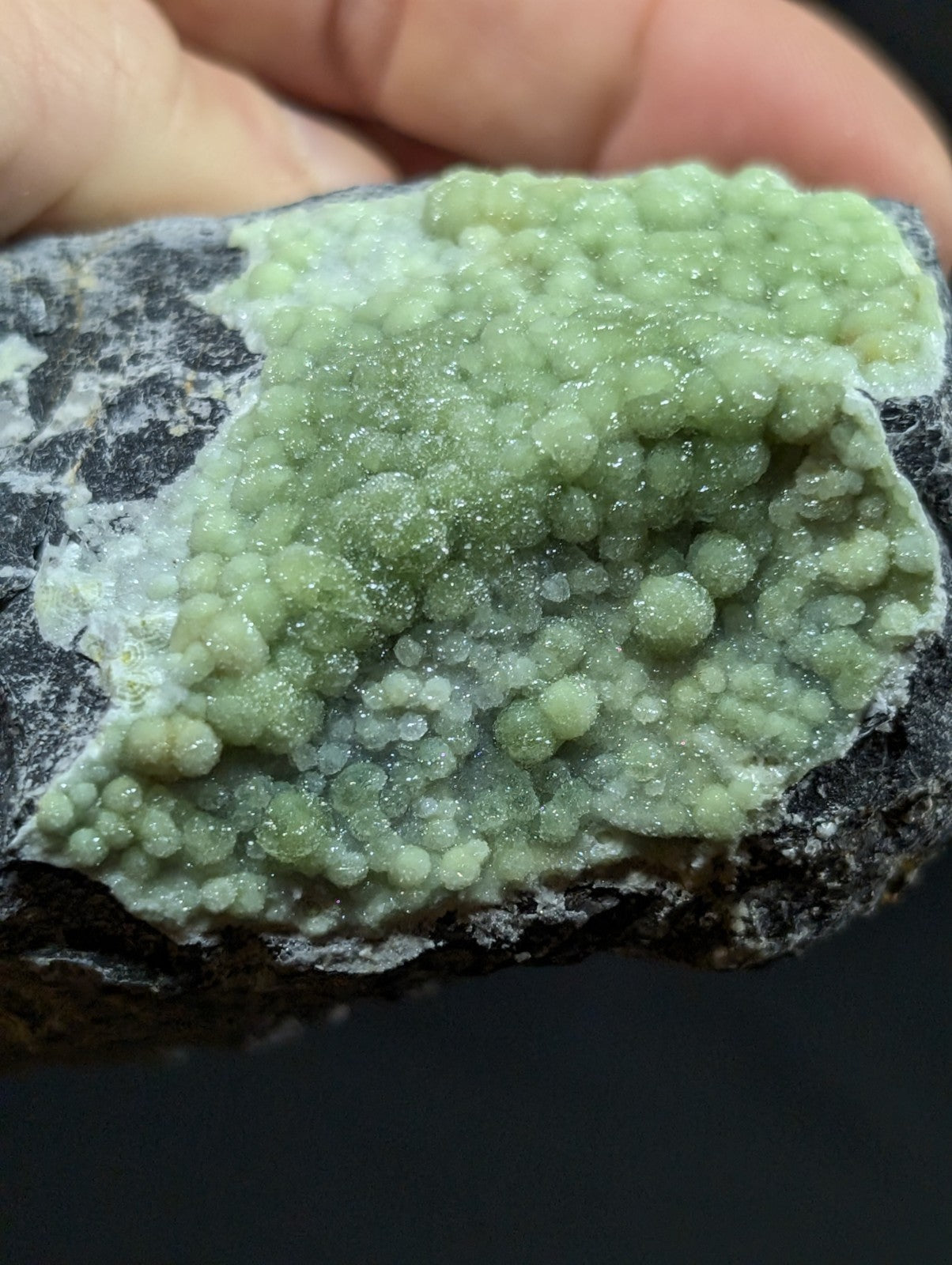 A hand holds a dark rock featuring a vibrant, light green crystalline formation with numerous small, rounded, glittering spheres clustered together.