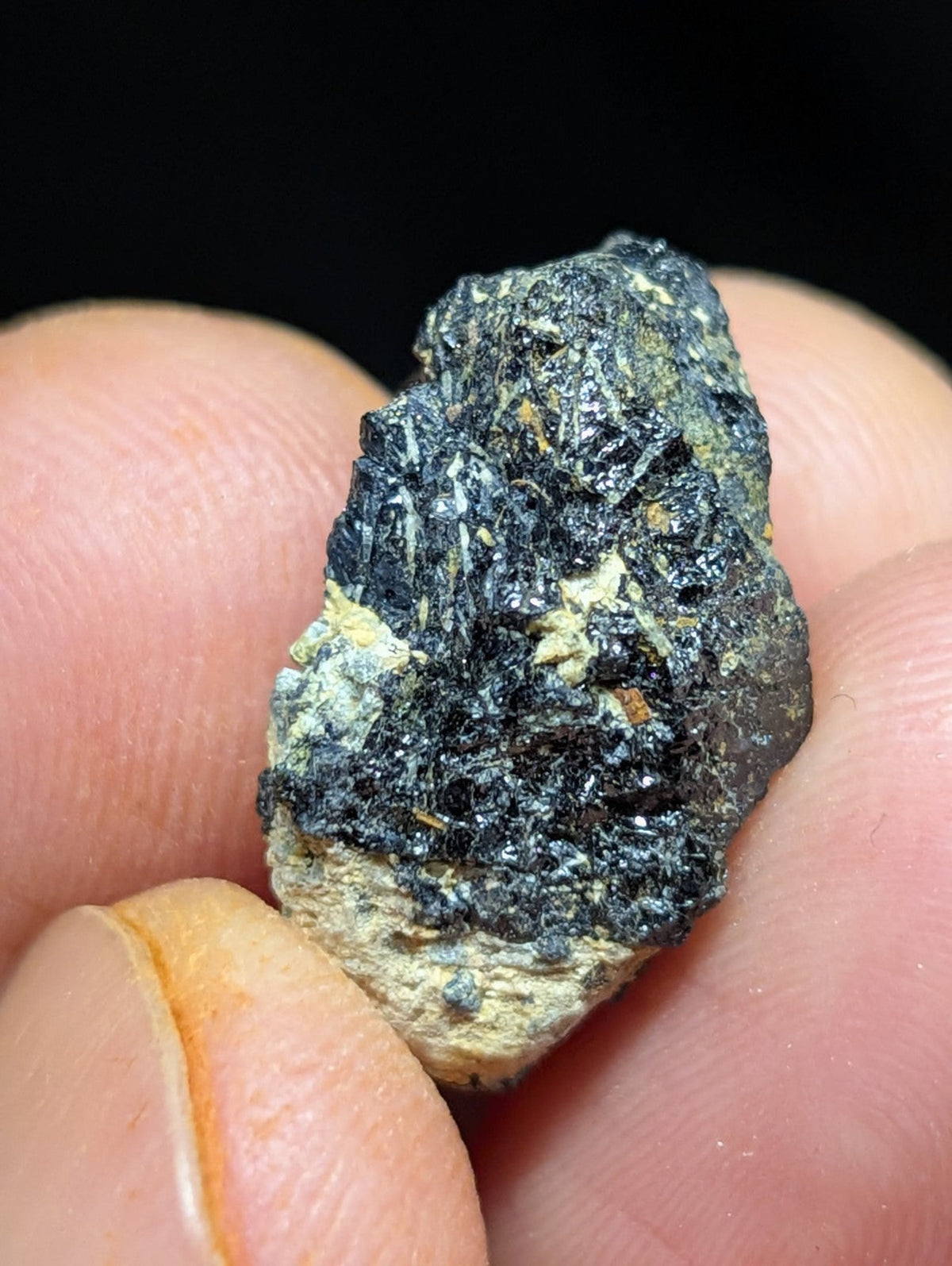 A rough, irregularly shaped mineral specimen with a dark black and greenish-gray surface texture held between fingers.