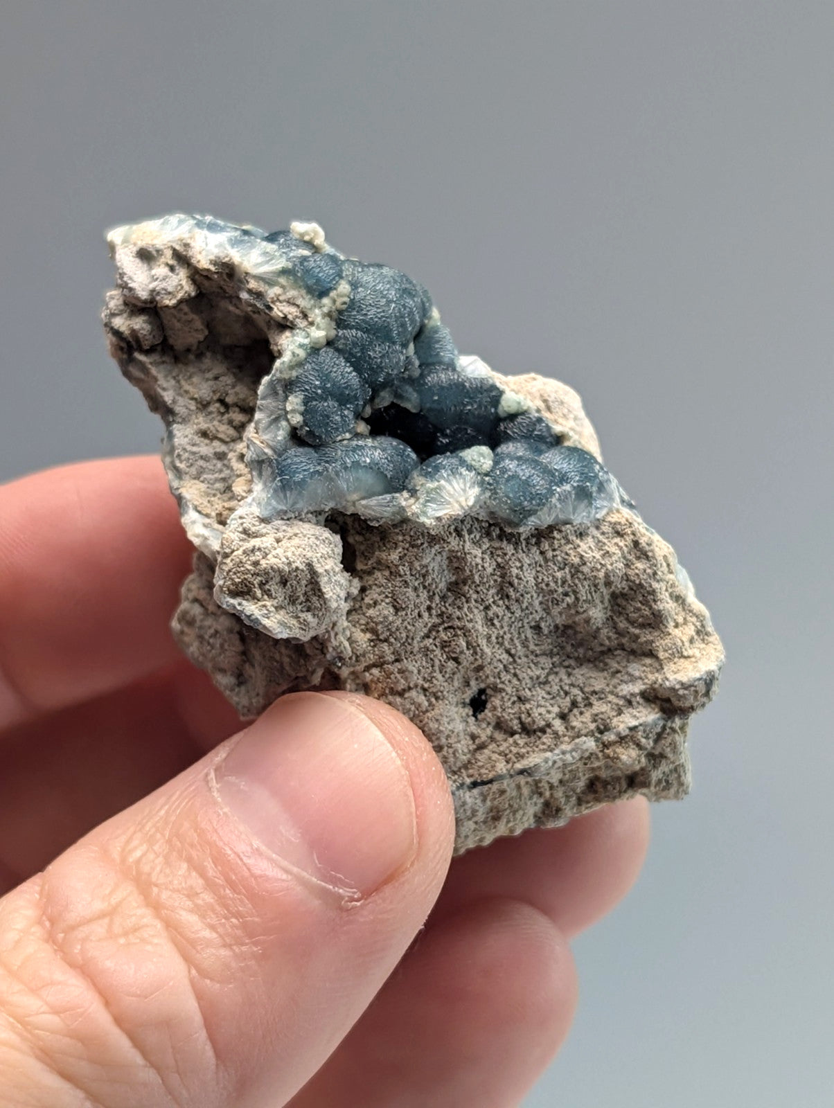 A hand holds a geological specimen featuring a cluster of translucent, deep blue druzy crystals emerging from a rough, light gray rock matrix.